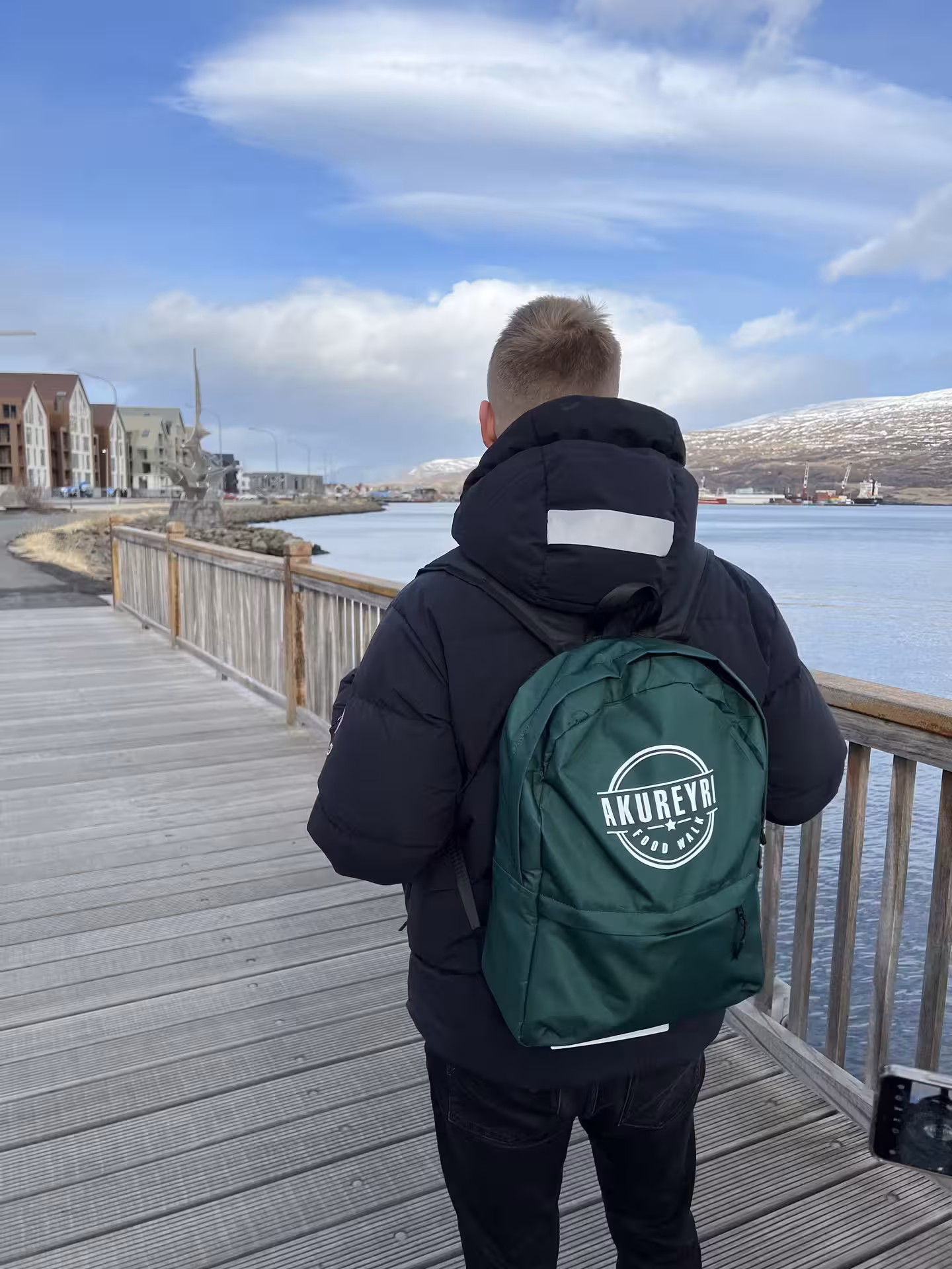 Person wearing Akureyri Food Walk green backpack on waterfront boardwalk with fjord and snowy mountains