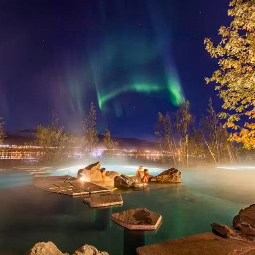 Northern lights glowing over steaming Forest Lagoon geothermal pool with rocky islands at night in Akureyri