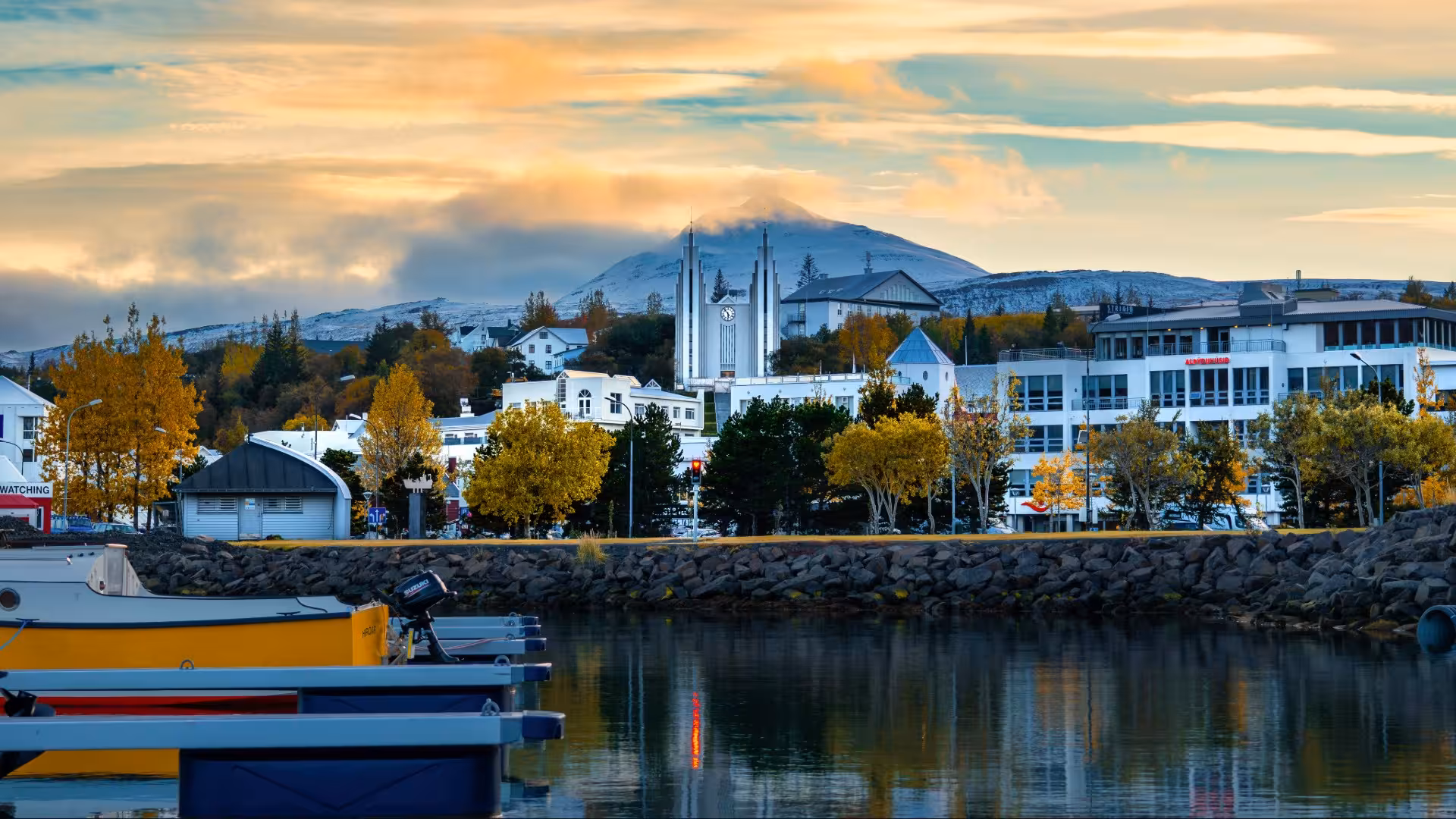 Akureyri harbor waterfront with Akureyrarkirkja church spires, autumn trees, and snow-capped mountain at sunset