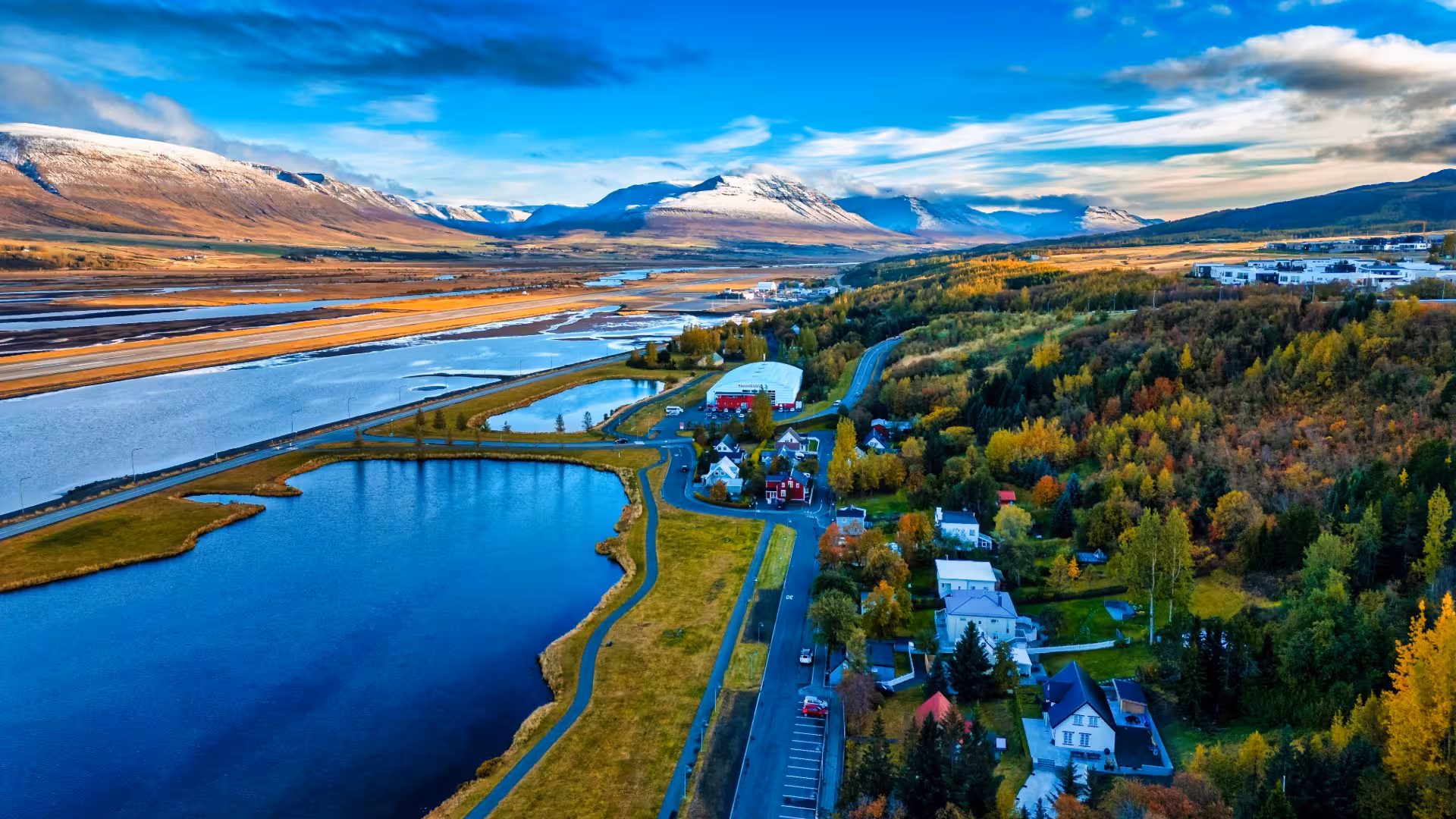 Aerial view of Akureyri town with lake, colorful autumn trees, road, and snow-capped mountains in Iceland