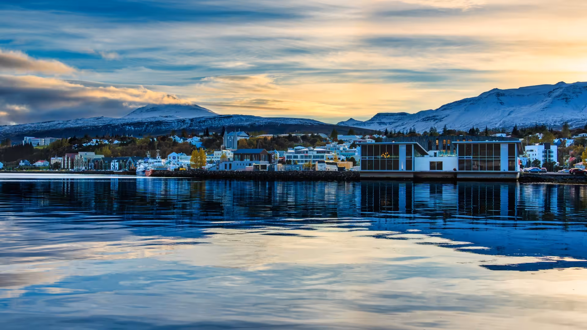 Akureyri harbor waterfront with colorful buildings, snow-capped mountains, and calm reflective water at sunset