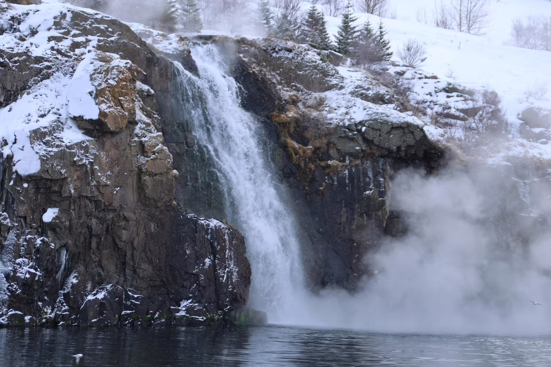Waterfall cascading over snow-covered rocky cliffs into steaming water in Akureyri, Iceland in winter