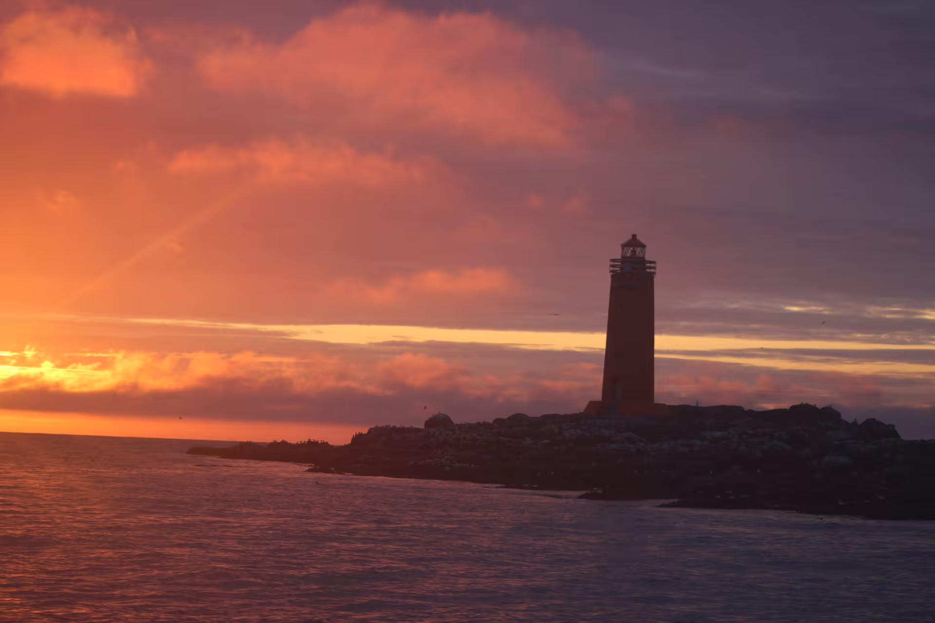 Lighthouse silhouette on rocky coast at sunset with vivid orange and purple sky over calm ocean waters