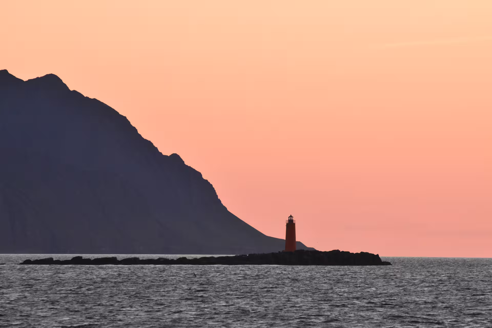 Red lighthouse on rocky islet at midnight sun with mountain silhouette and orange sky in Akureyri