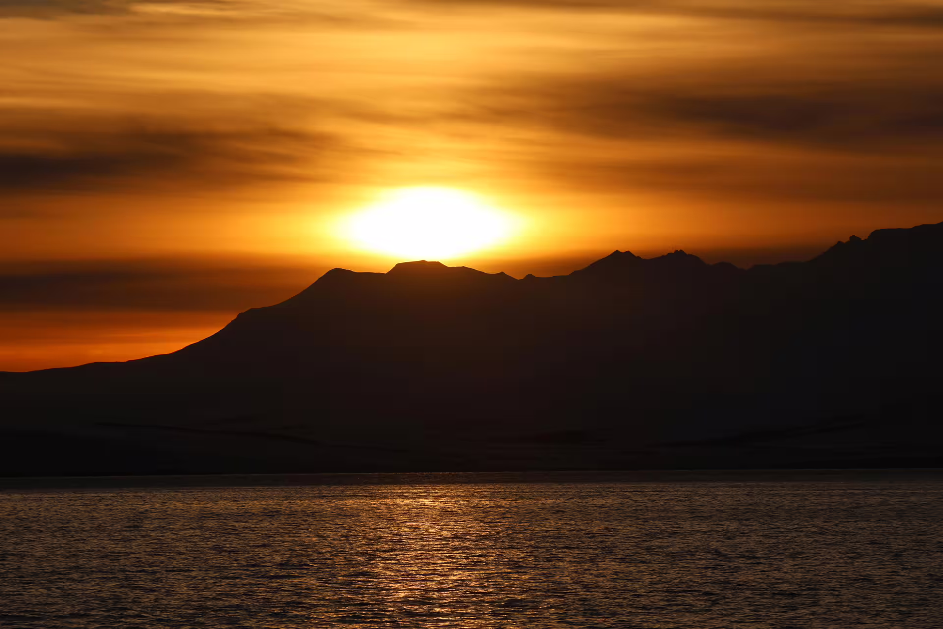Midnight sun setting over dark mountain silhouettes reflected on calm Arctic ocean waters in Akureyri