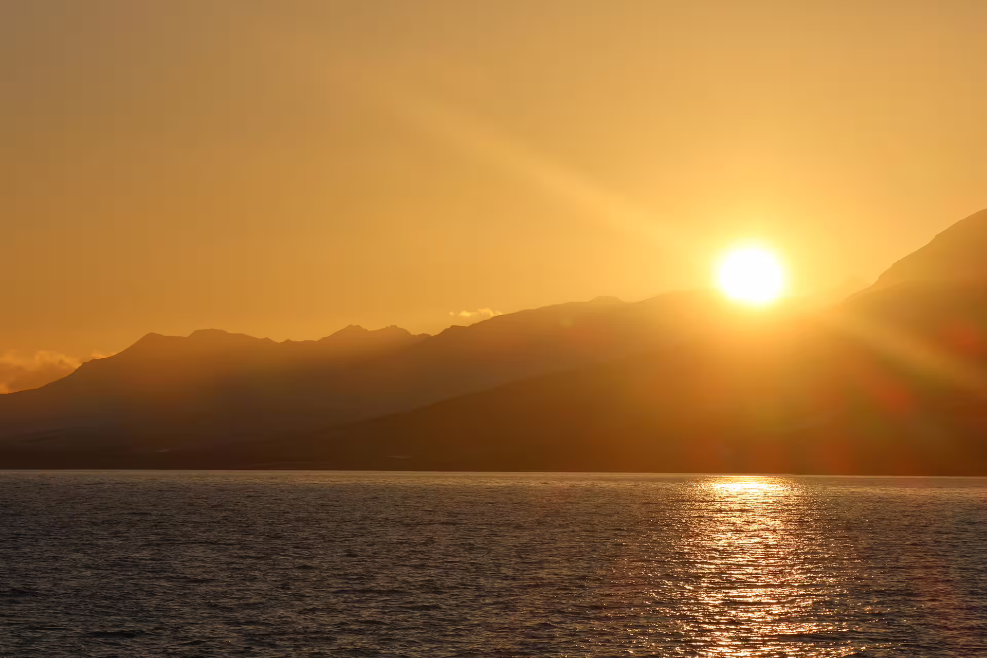 Golden midnight sun setting over mountain silhouettes reflected on calm ocean waters in Akureyri