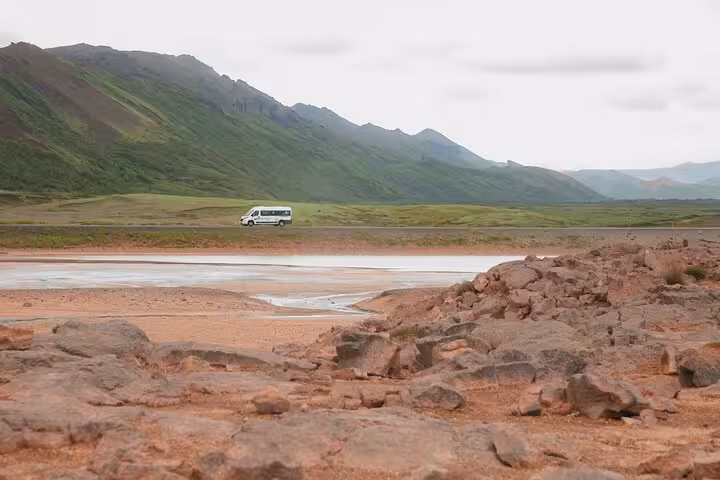 White tour bus driving through green Icelandic mountain valley with rocky terrain in foreground