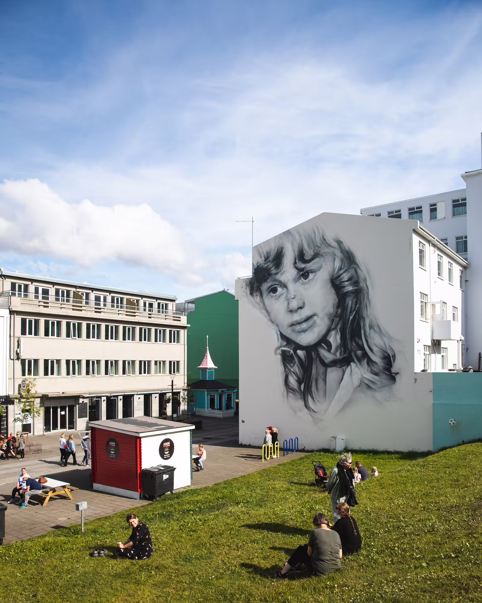 Large black and white portrait mural on building wall in Akureyri town square with people relaxing on grass