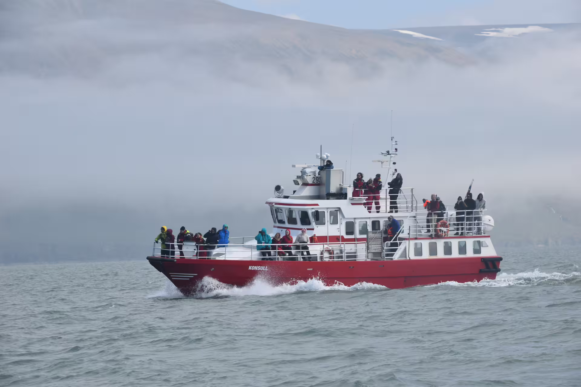 Red and white Konsull whale watching boat filled with tourists sailing through misty Eyjafjordur Fjord near Akureyri