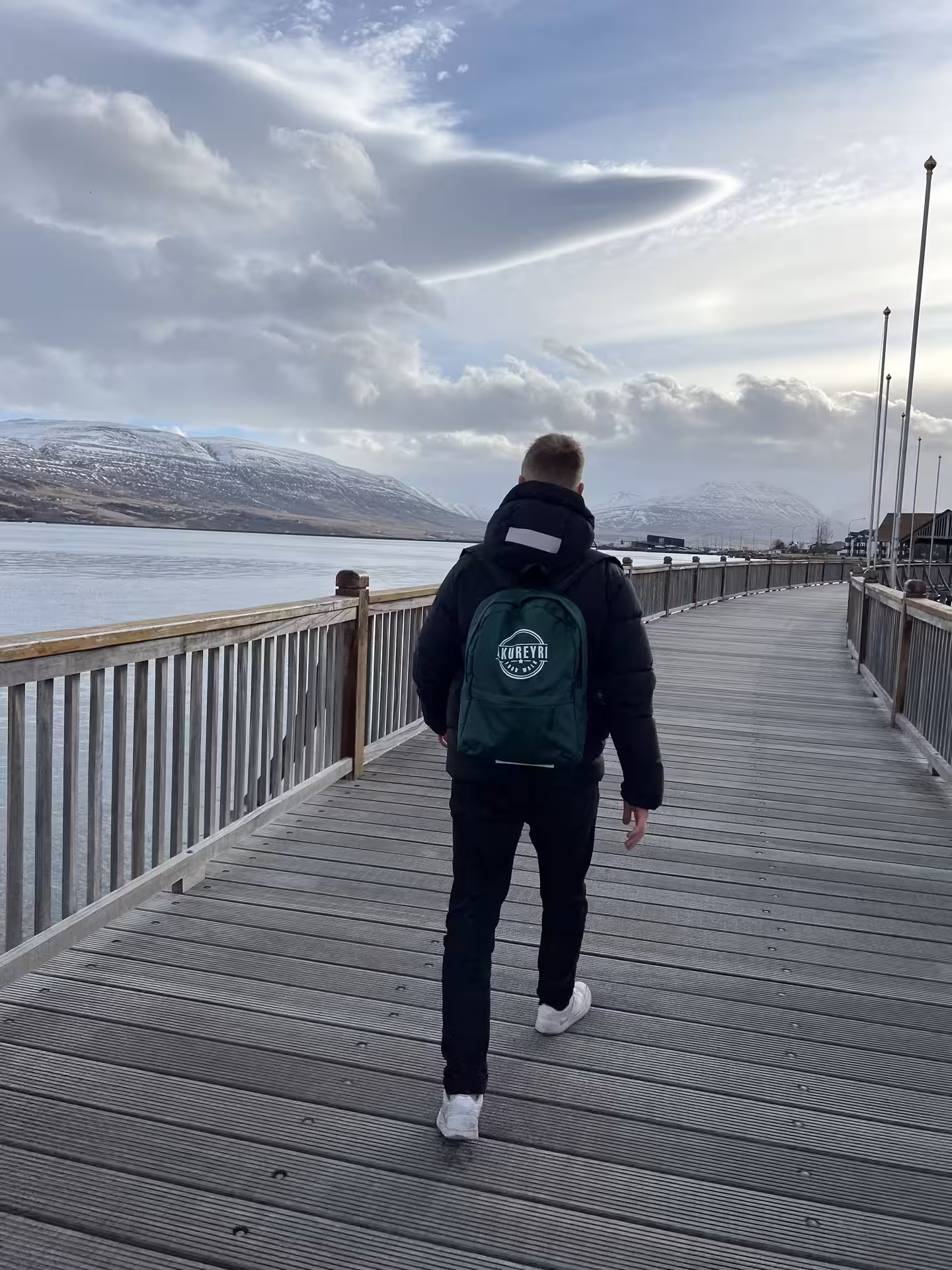 Person with Akureyri backpack walking wooden pier overlooking snowy mountains and fjord in Iceland
