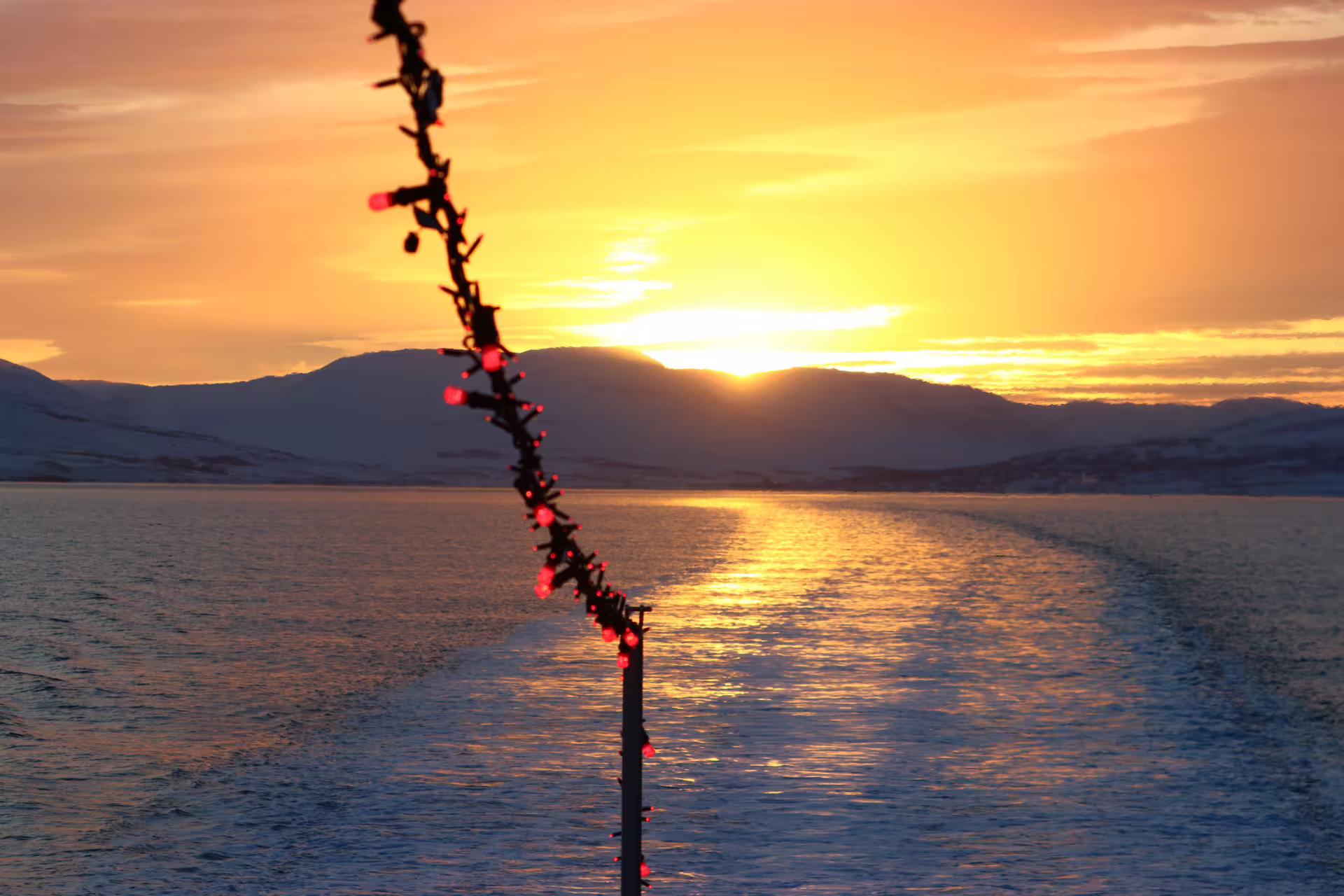 Red string lights on a ship mast with golden midnight sun setting over mountains and calm fjord waters in Akureyri