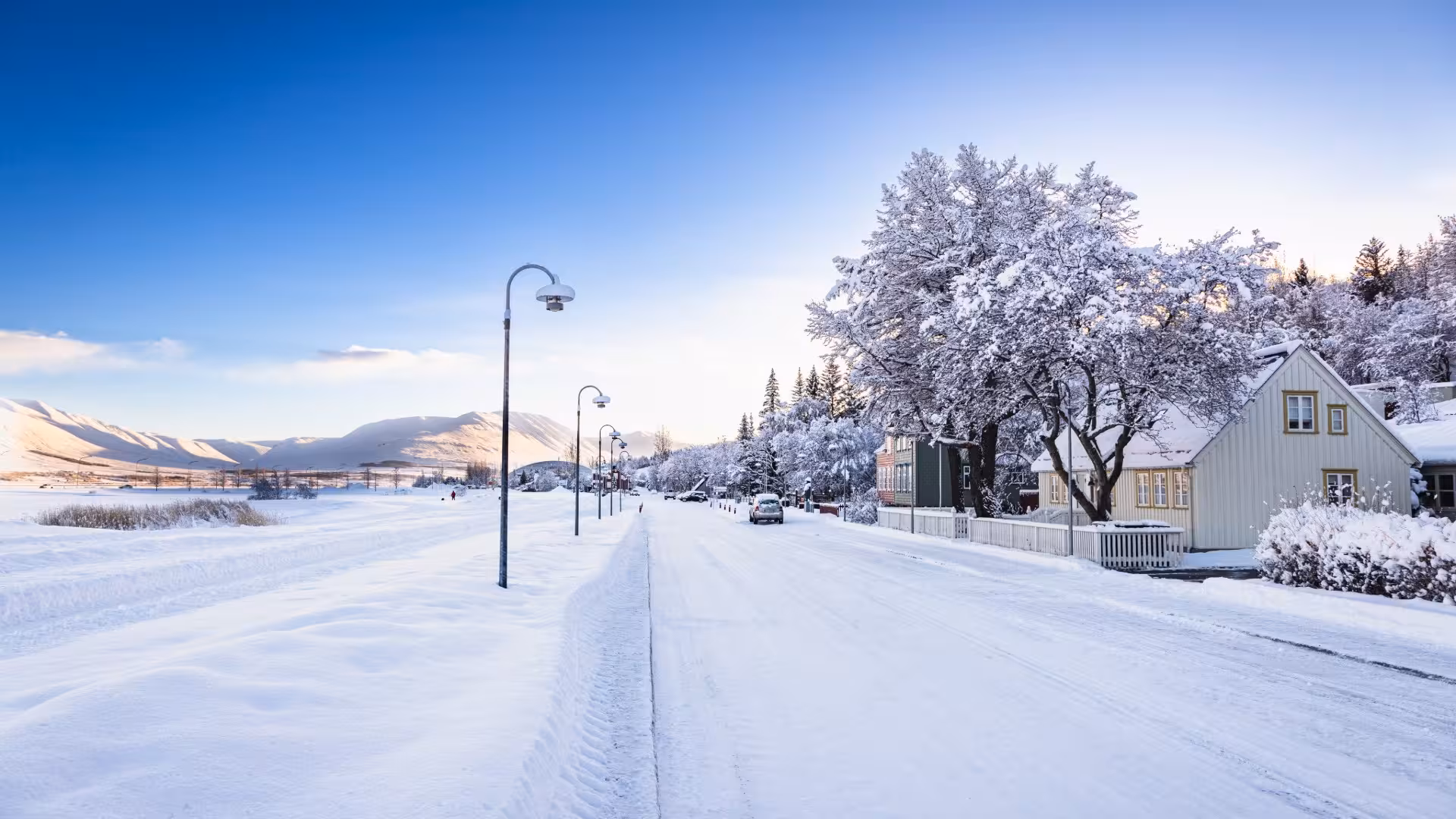 Snow-covered street in Akureyri with traditional houses, lamp posts, and mountains in winter