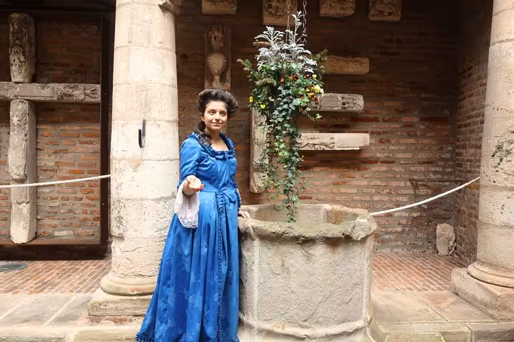 Guide in blue 18th-century dress beside a stone well in Albi's historic courtyard