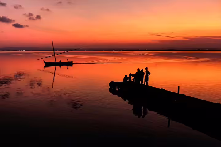 Silhouetted figures enjoy a serene sunset boat ride on Albufera Lagoon, highlighting a private day trip with paella and nature trek.