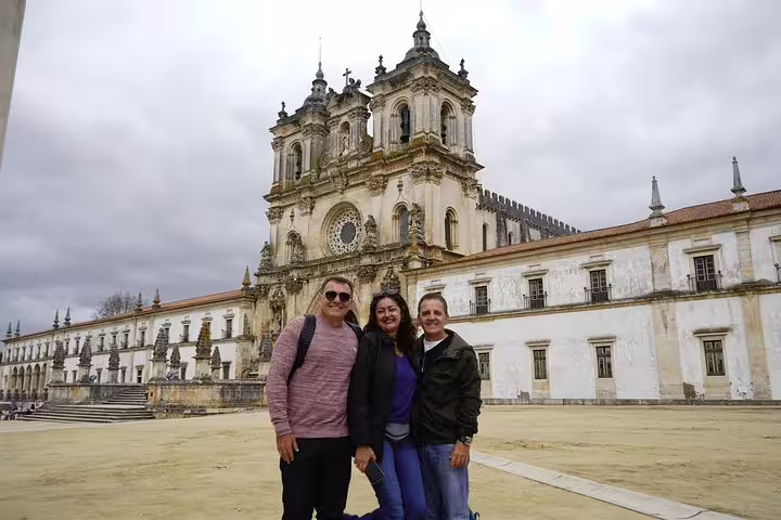 Three tourists smiling in front of the historic Alcobaça Monastery during a private full-day tour from Lisbon to Óbidos, Nazaré, Batalha, and Fátima.