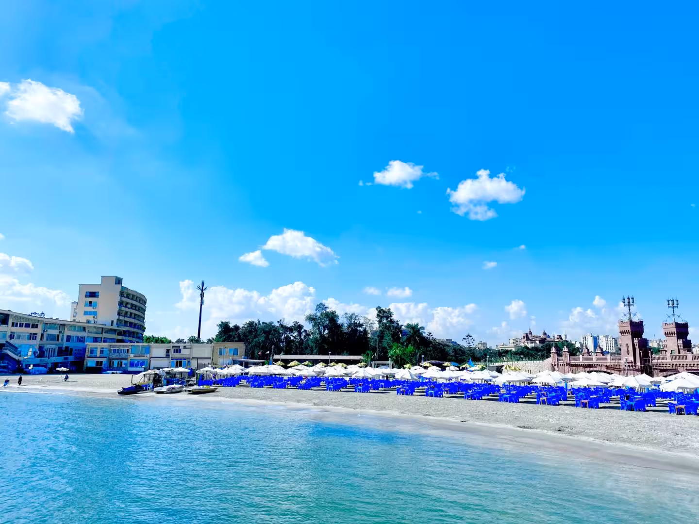 Alexandria beach with blue chairs, white umbrellas, and a castle tower along the Mediterranean shore