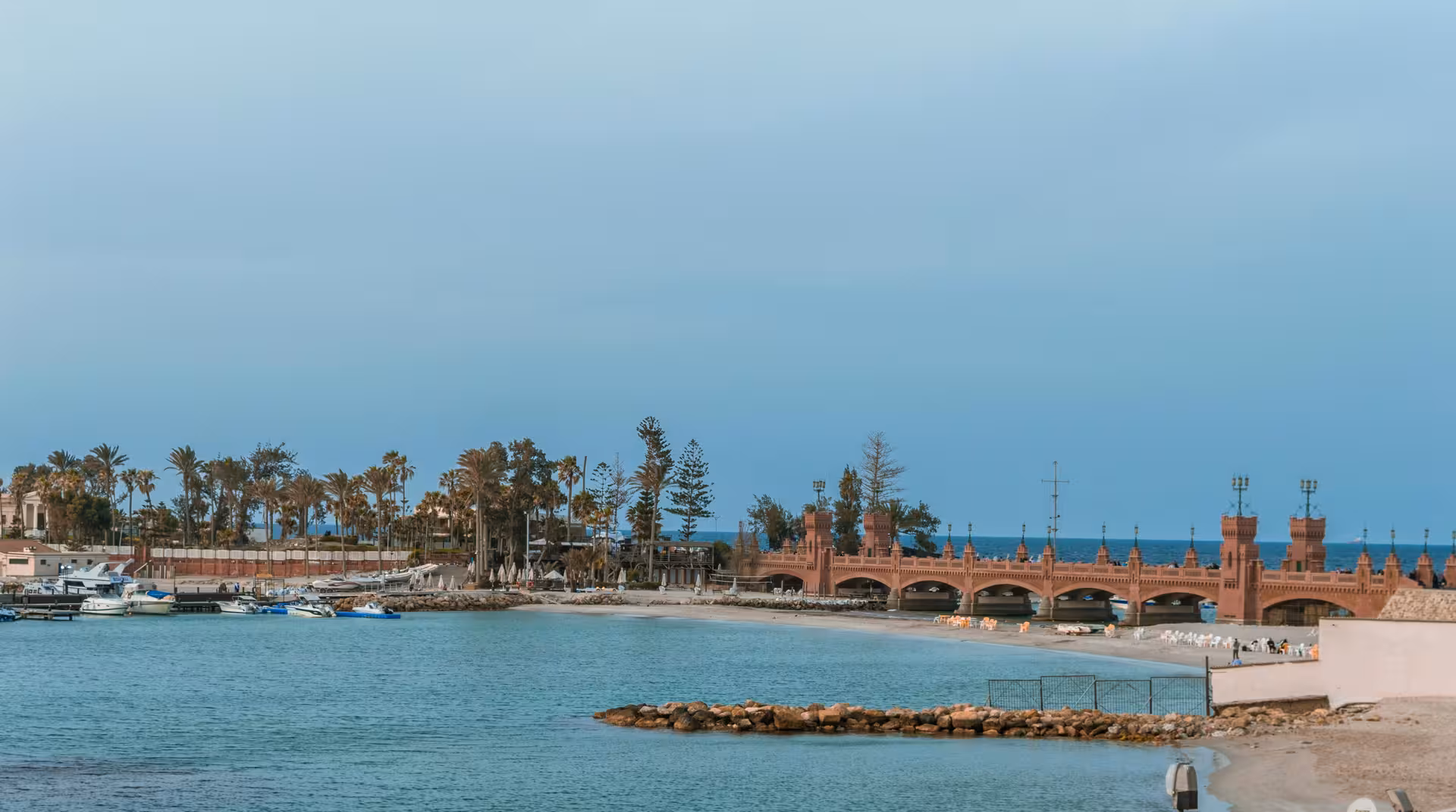 Alexandria waterfront with ornate red brick bridge, palm trees, marina boats, and calm blue Mediterranean sea