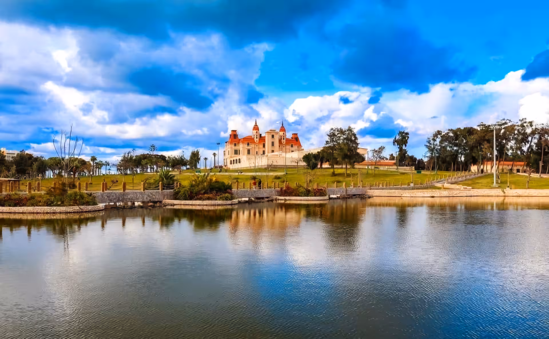Historic palace with red rooftops reflected in a calm lake surrounded by green parkland in Alexandria, Egypt