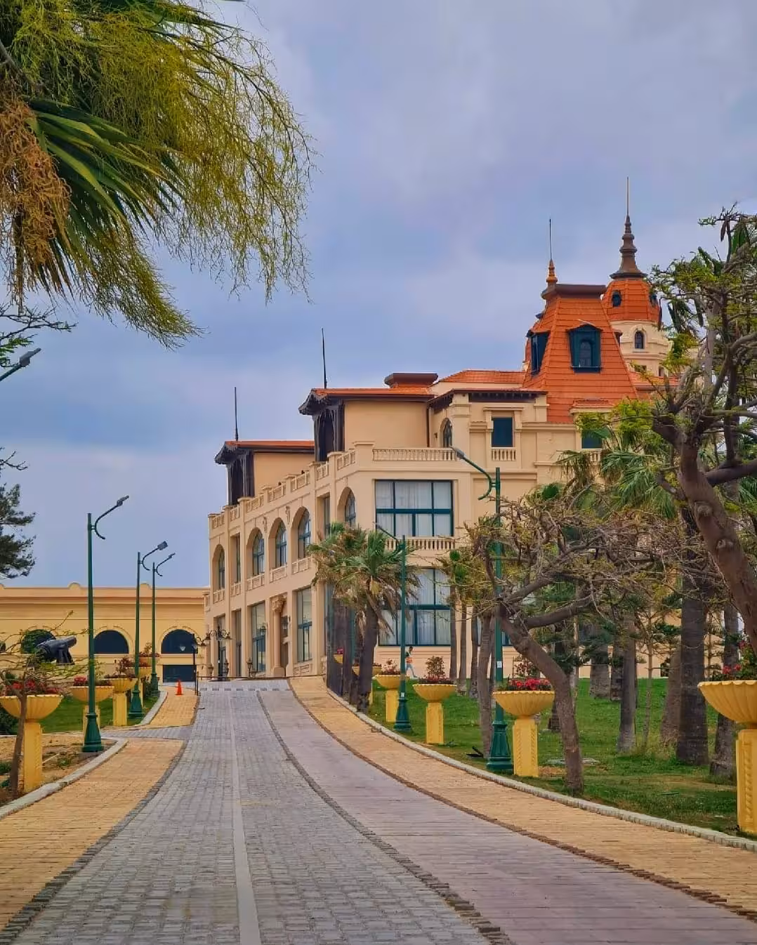 Tree-lined promenade leading to a grand historic palace with terracotta roof in Alexandria Royal Gardens