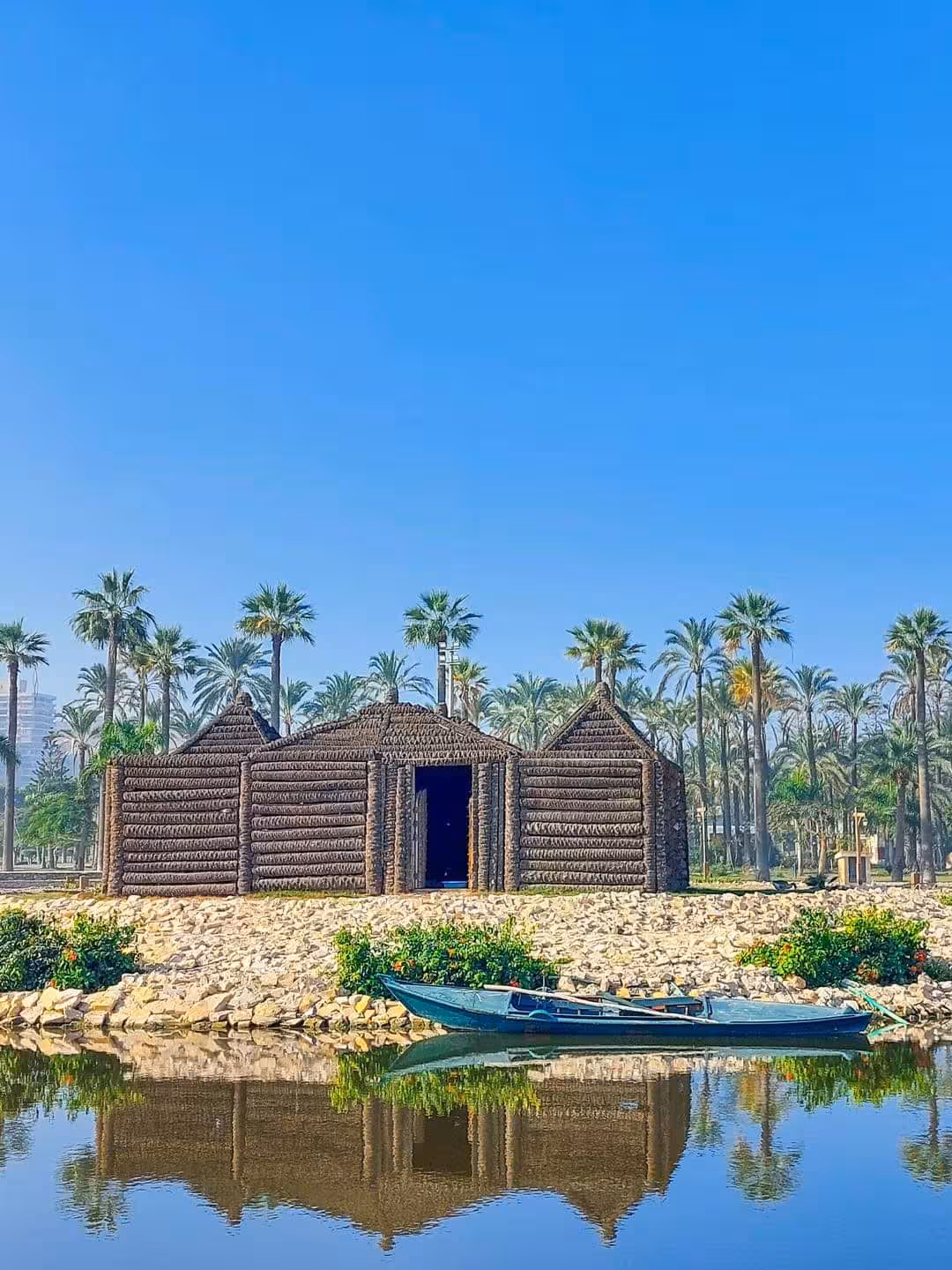 Traditional reed huts on Alexandria waterfront with palm trees and blue rowing boat reflected in calm water