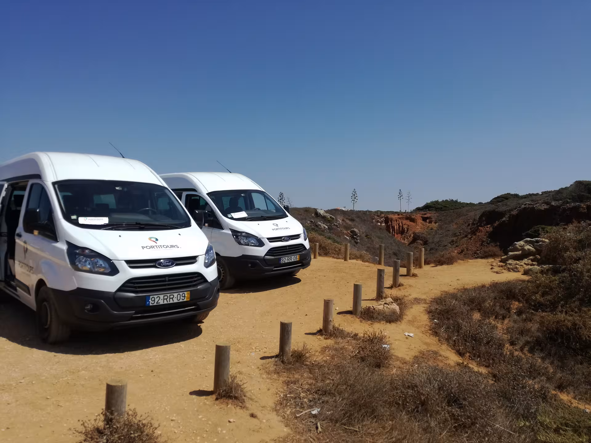 Comfortable Algarve à la Carte tour minibuses parked by dramatic coastal cliffs under clear blue skies in southern Portugal