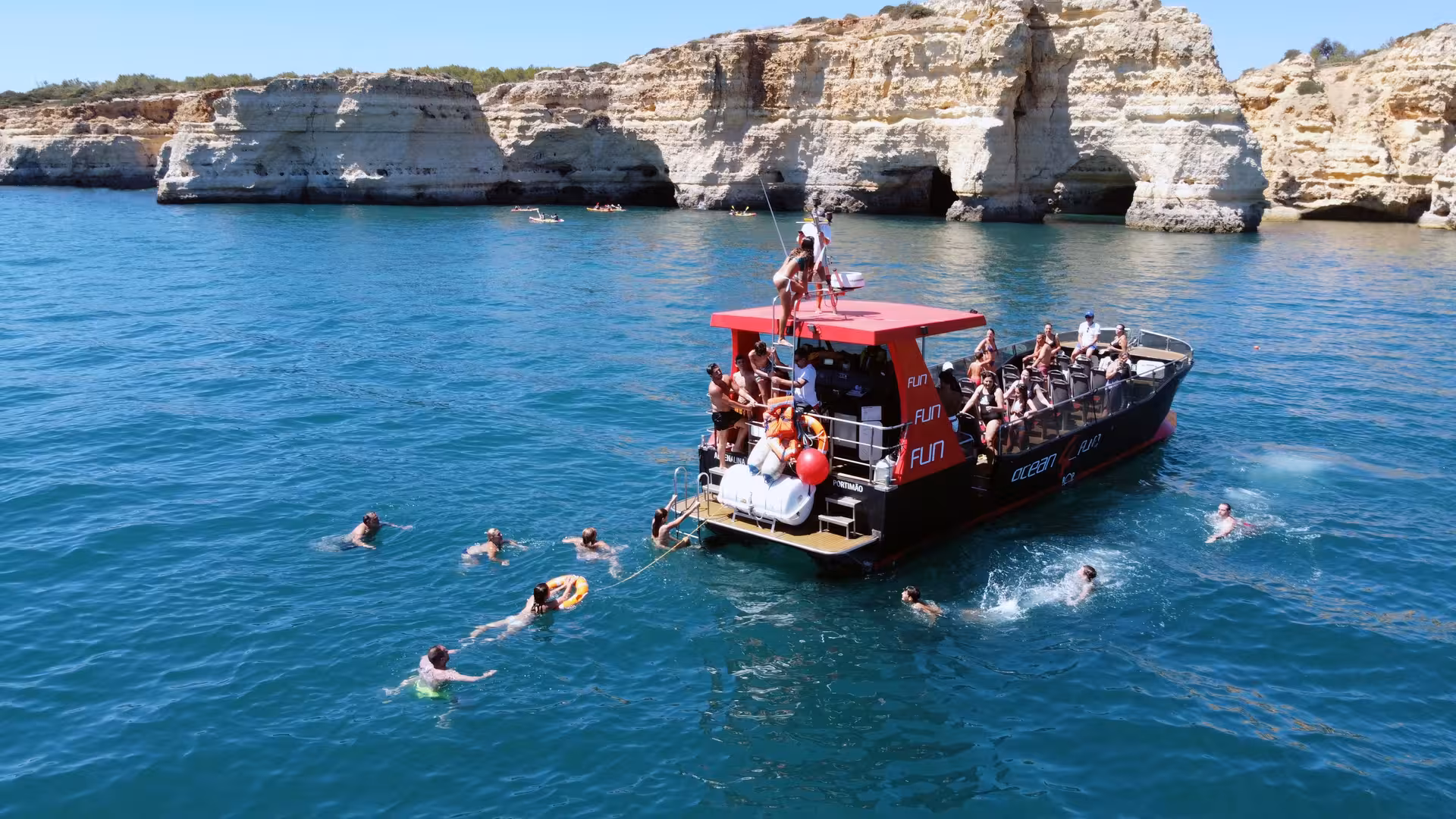 Guests swimming in turquoise waters beside a tour boat near dramatic Algarve limestone cliffs and sea caves on a Portimao coastal cruise