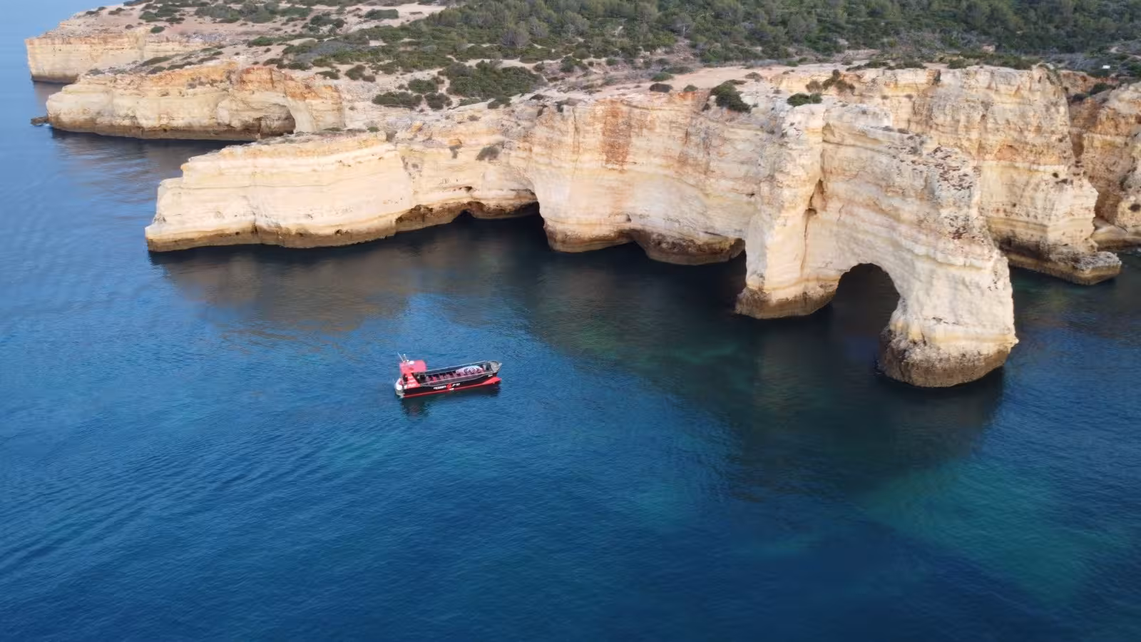 Aerial view of a red tour boat sailing near stunning golden limestone sea caves and arches along the Algarve coast from Portimao