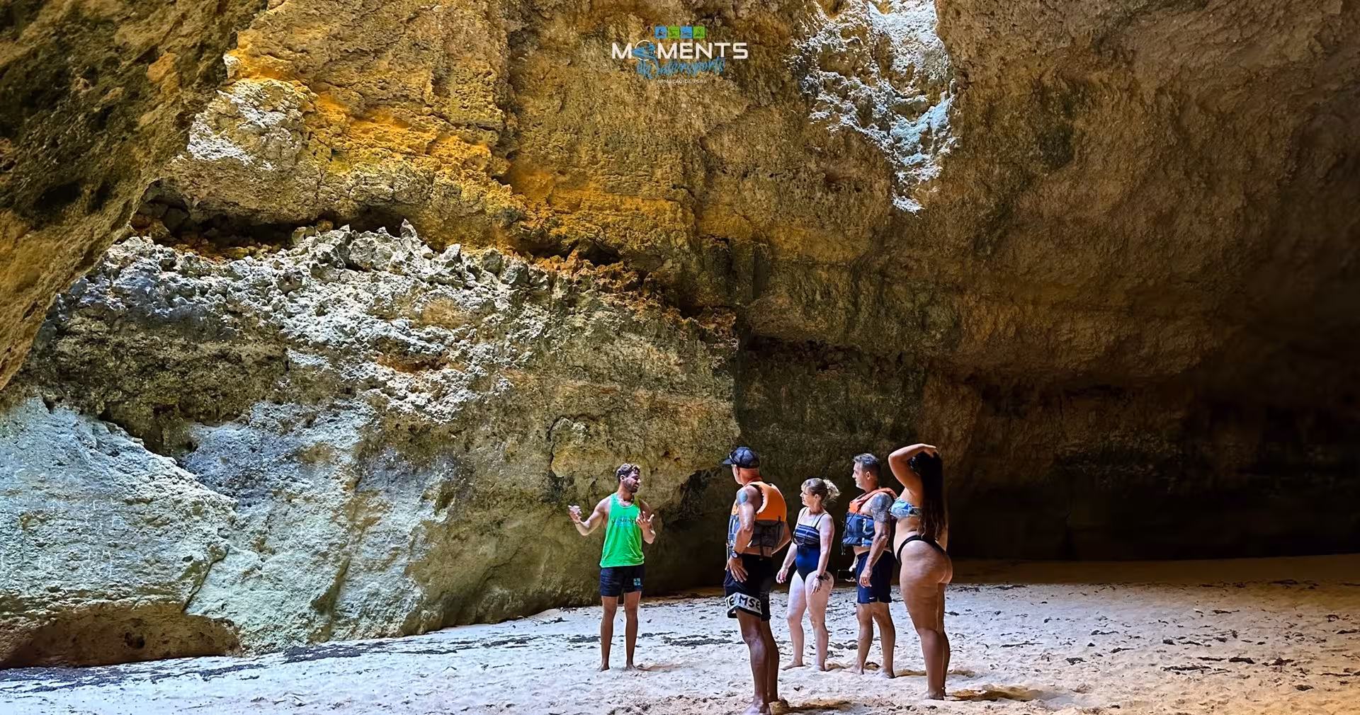 A tour guide explains the stunning interior of an Algarve sea cave to a small group on a sandy floor during a kayak adventure tour
