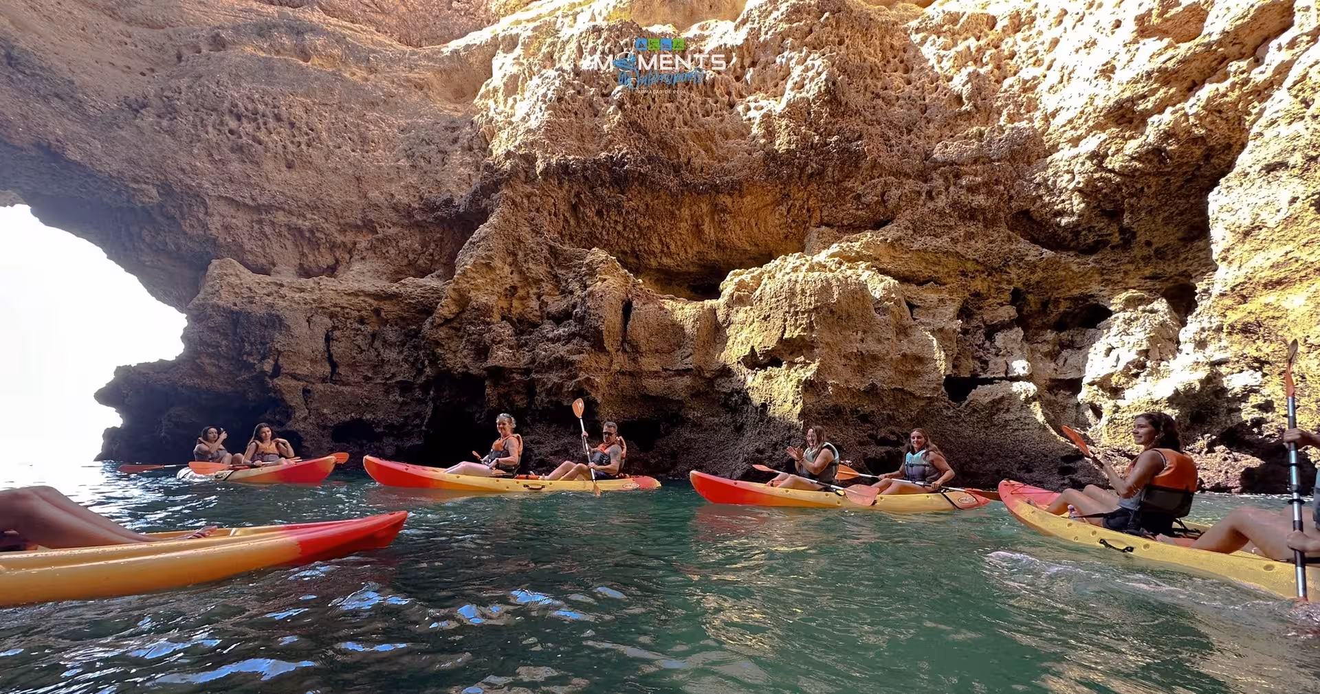 Group of kayakers paddling in colorful sit-on-top kayaks beside dramatic golden limestone sea caves on the Algarve coastline Portugal