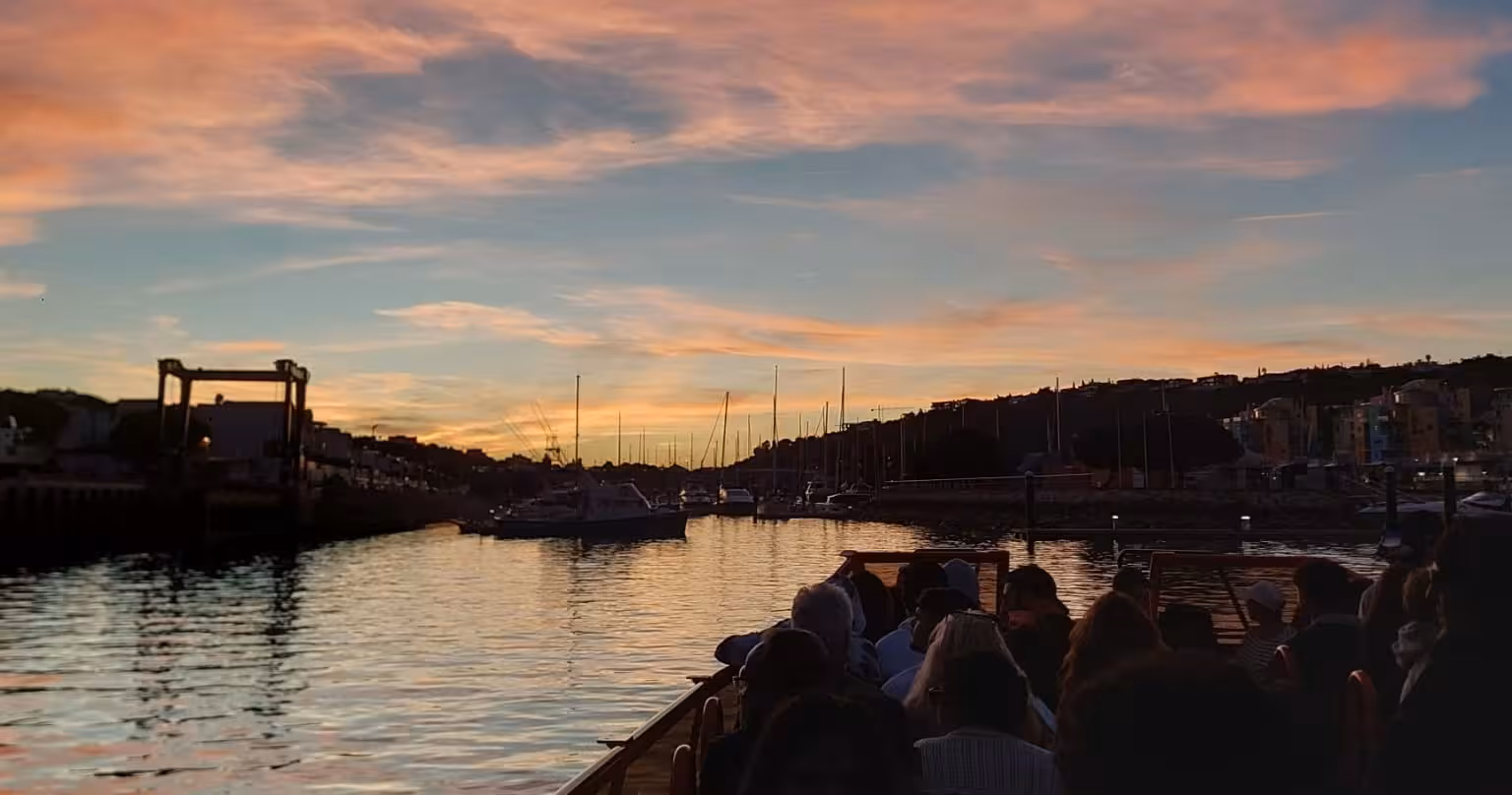 Crowd on open-top boat departs marina at dusk for Algarve sunset tour, passing sailboats under pastel pink and blue sky