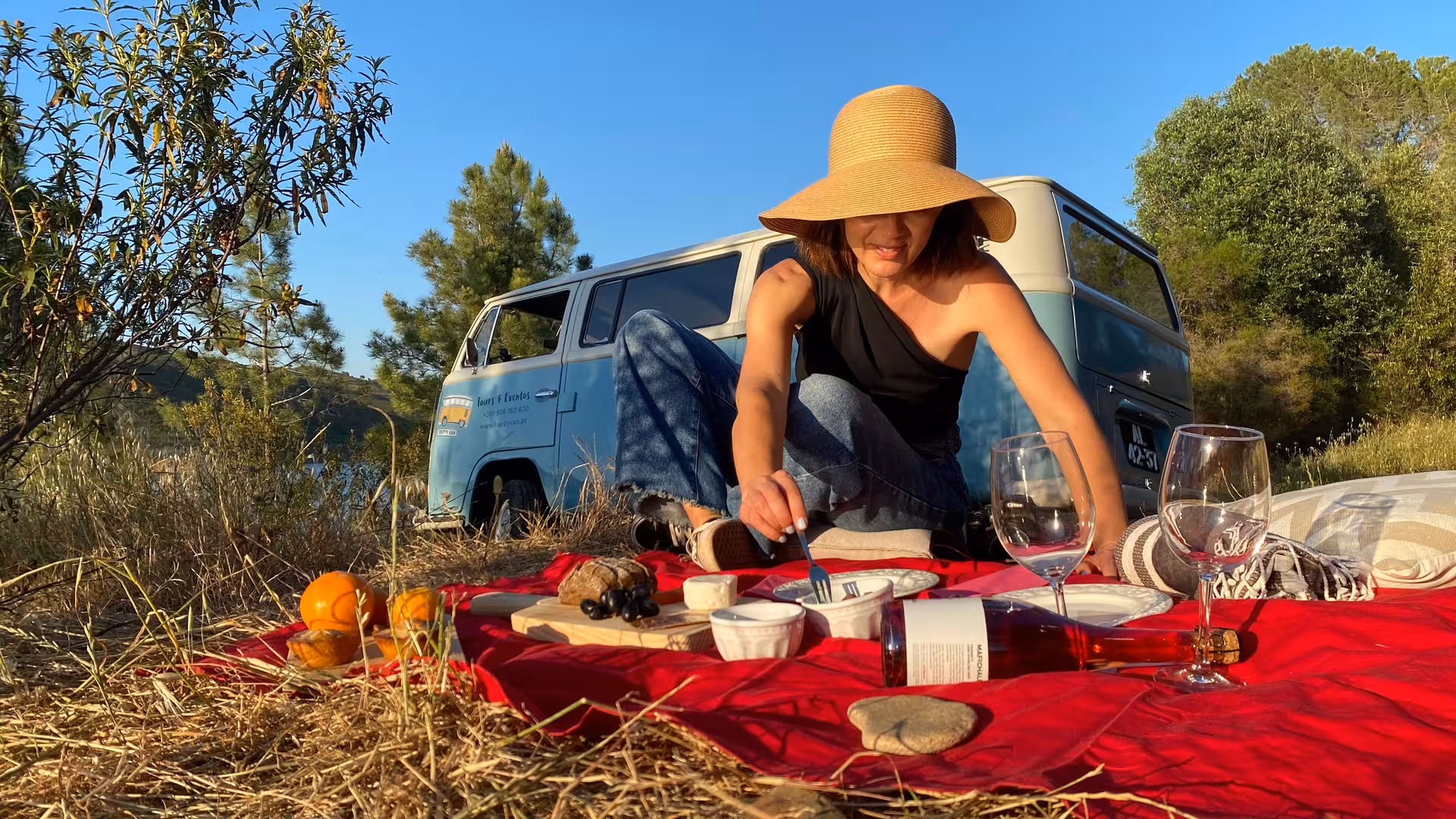 Woman enjoying gourmet picnic with wine and cheese by a vintage van in Algarve vineyard.
