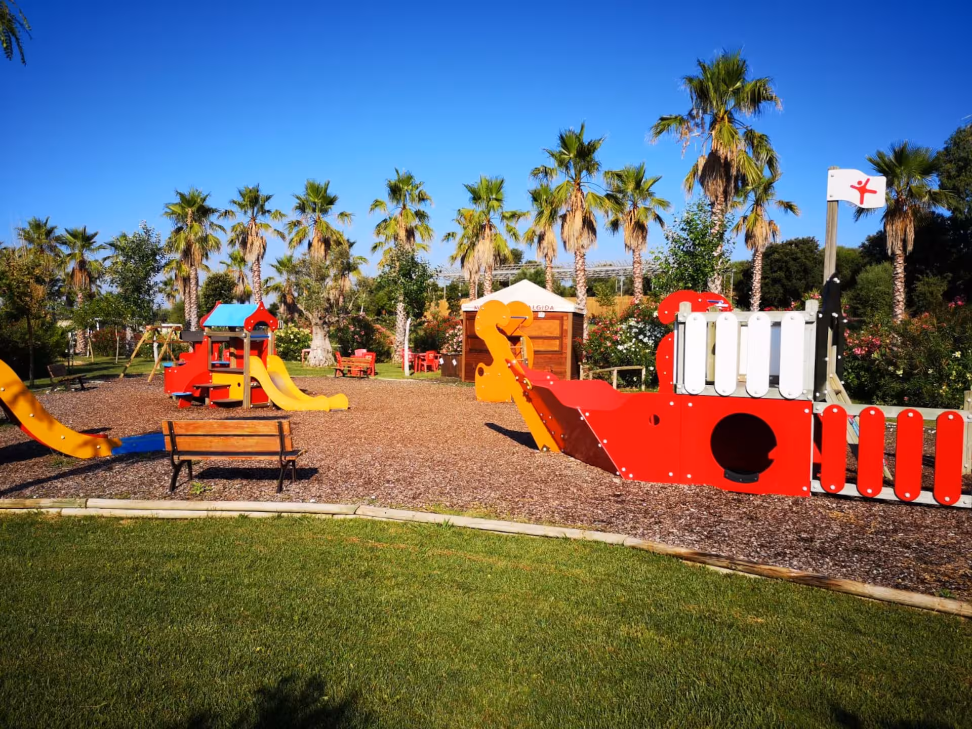 Colorful children's playground set amidst palm trees offers fun activities at Alghero Butterfly House visit.