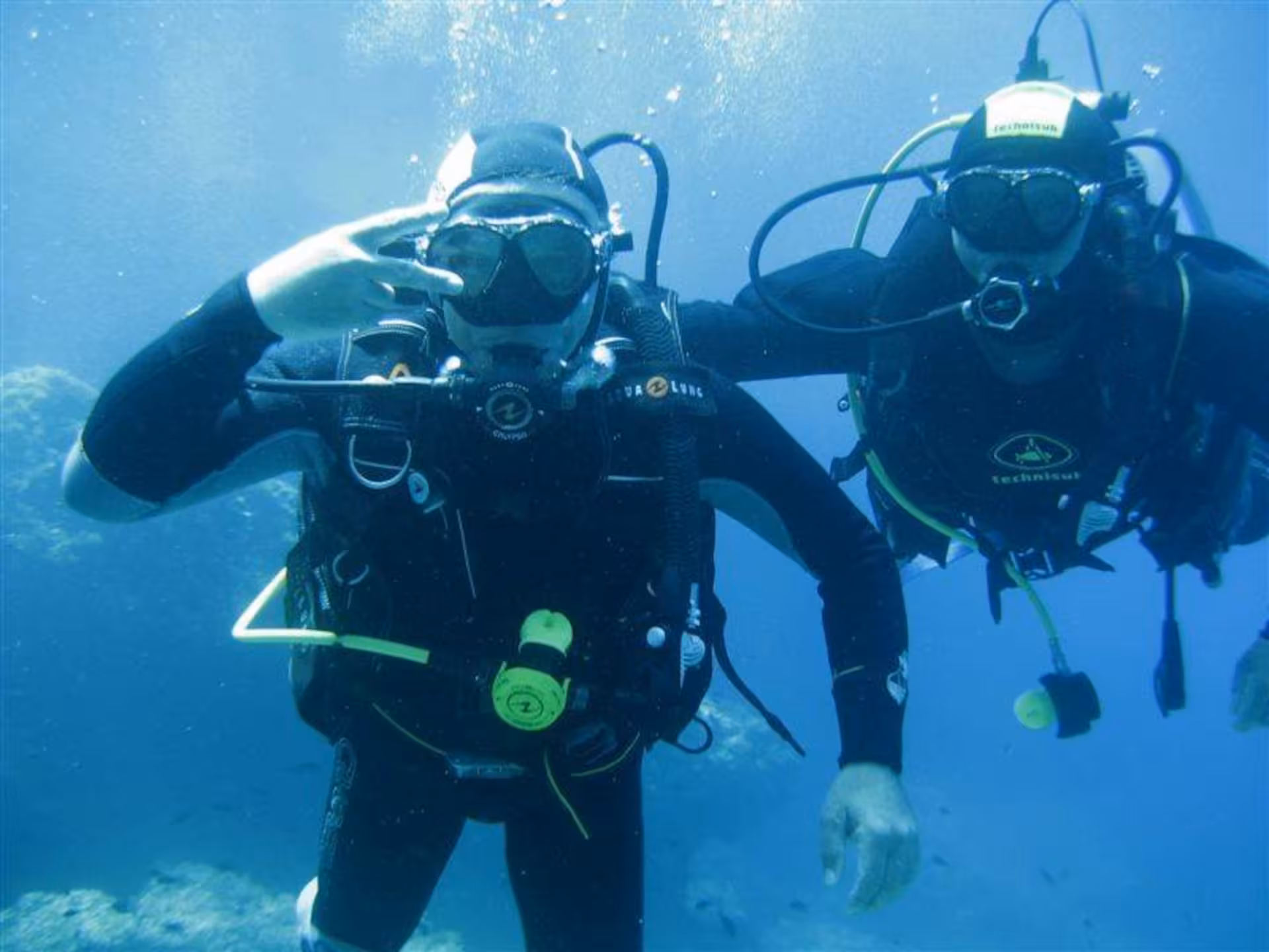 Two divers explore Capo Caccia's underwater marvels in Alghero, wearing full scuba gear in clear blue waters.