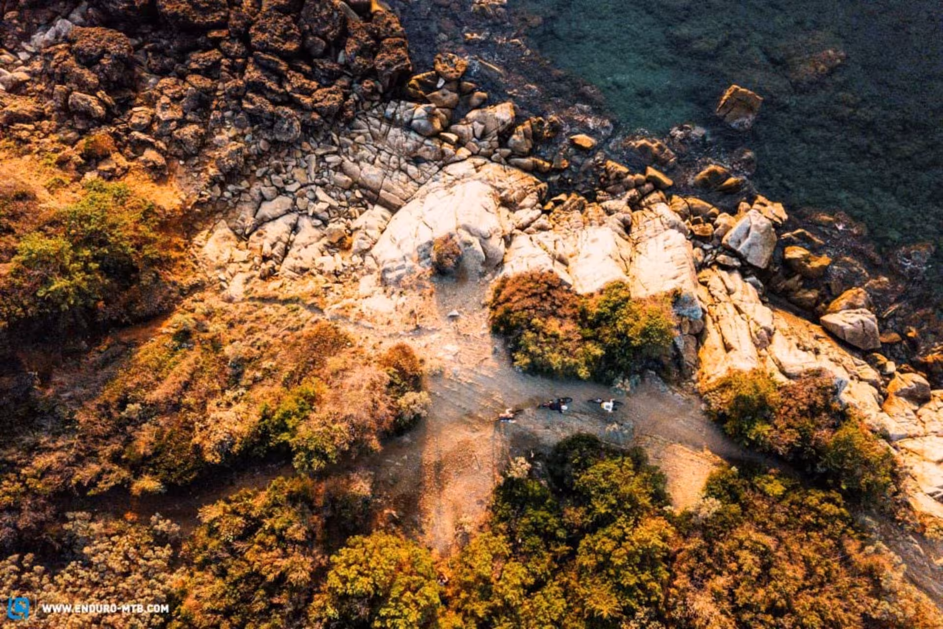 Aerial view of rugged Alghero coastline with cyclists on a scenic e-bike trail surrounded by rocks and greenery.