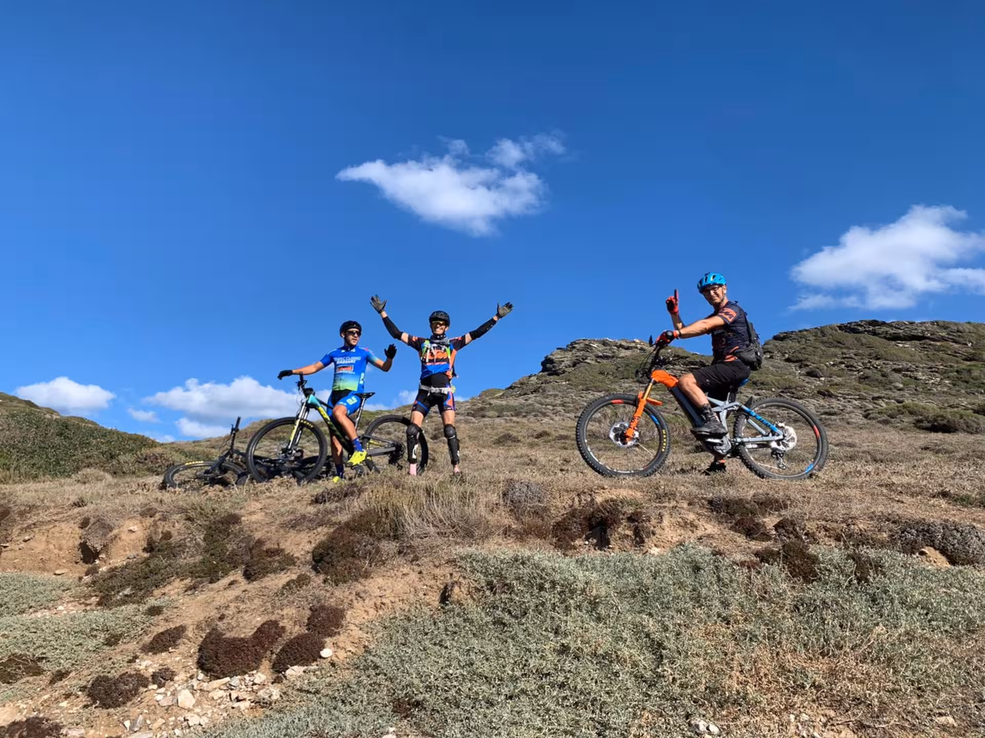 Three cyclists enjoying a sunny e-bike tour along the rugged Alghero coastline with clear blue skies in the background.