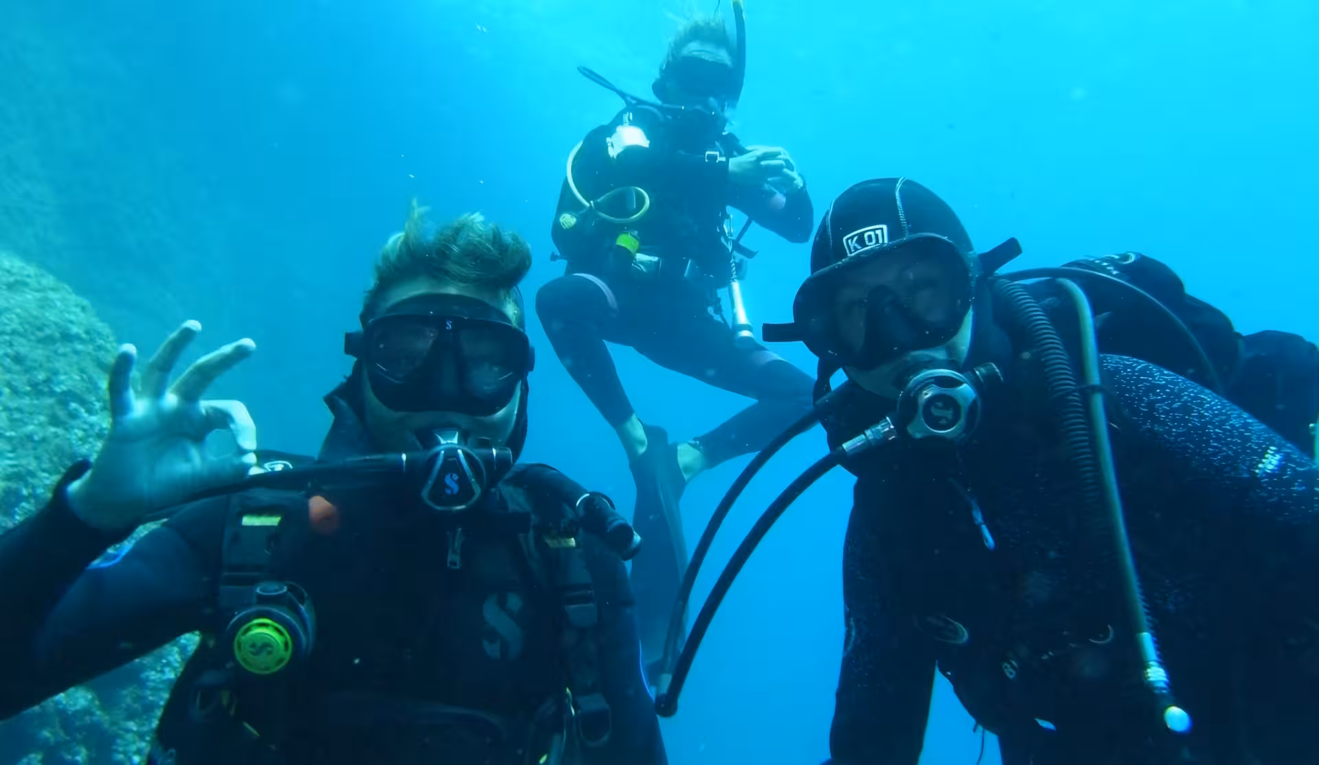 Three divers exploring underwater in Alghero, showcasing scuba gear and signaling OK during a diving baptism experience.