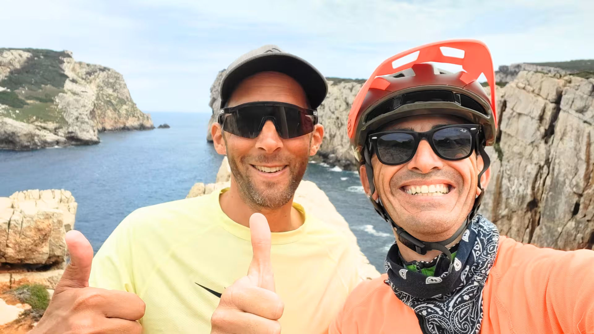 Two cyclists smiling with thumbs up at Capo Caccia cliffs during guided ebike tour in Alghero.
