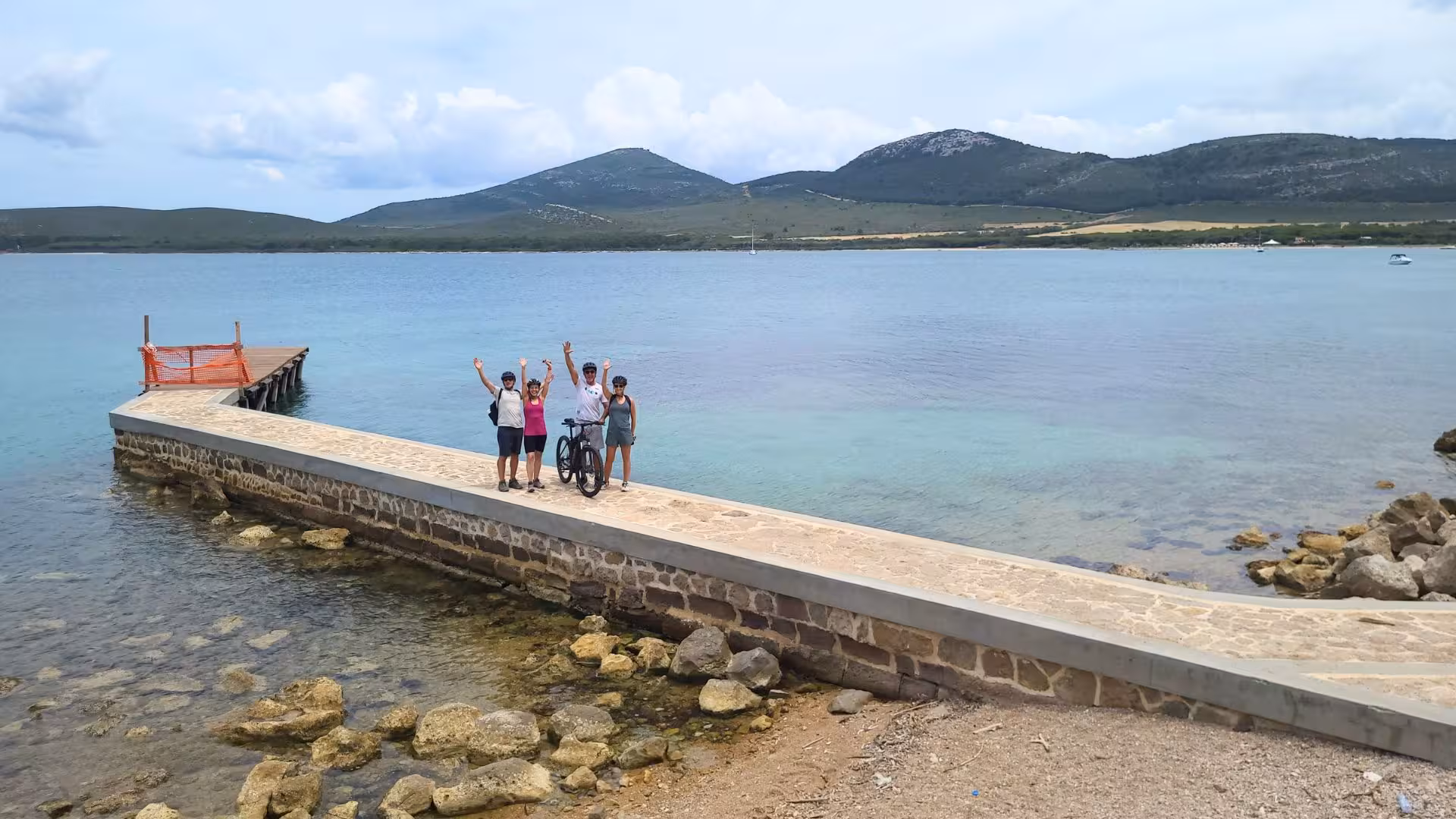 Group celebrates on a pier during the Alghero ebike tour, surrounded by the stunning turquoise waters of Capo Caccia.