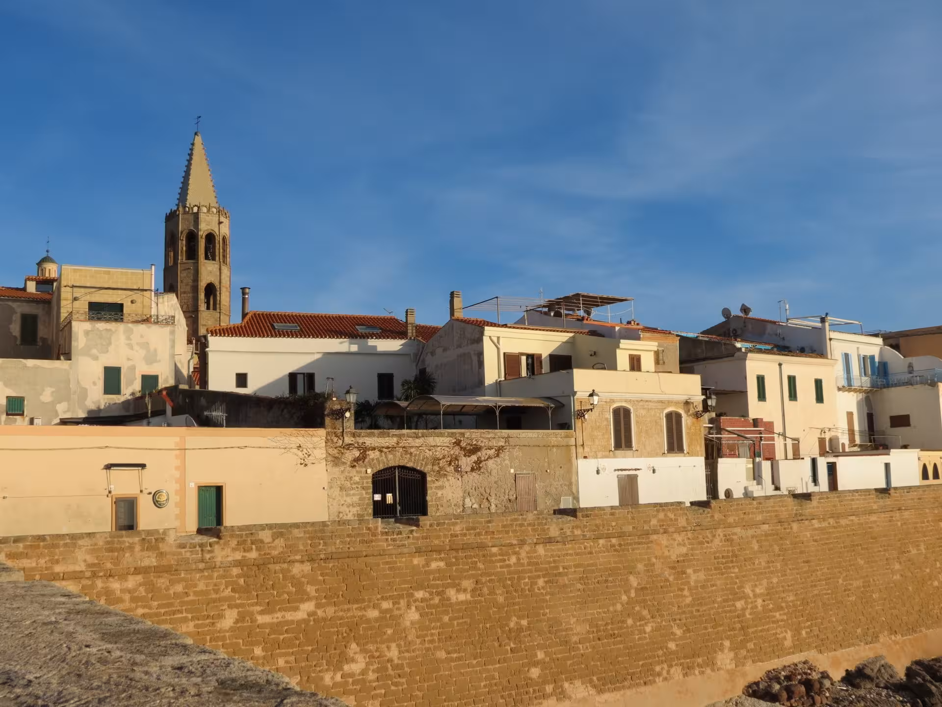 Scenic view of Alghero's historic center with charming buildings and a prominent bell tower at sunset.