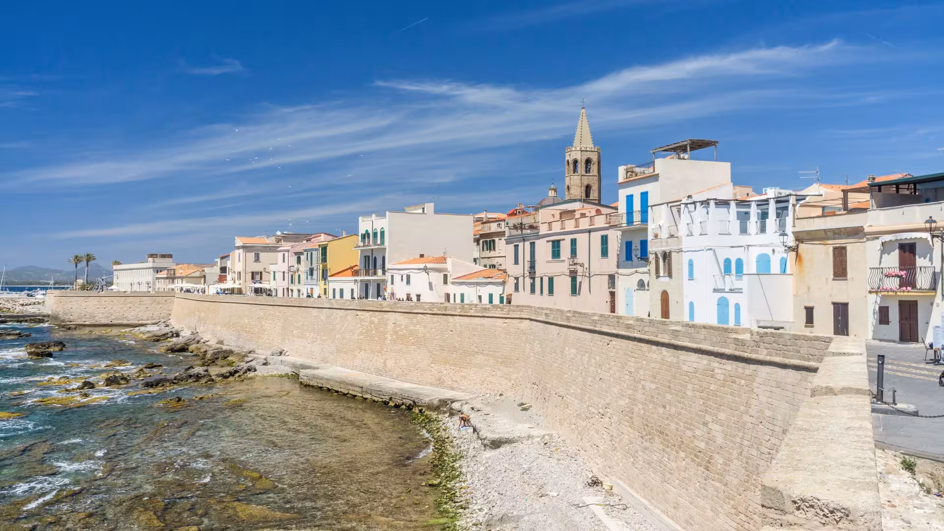Picturesque coastal view of Alghero's historic buildings along the city walls under clear blue skies.