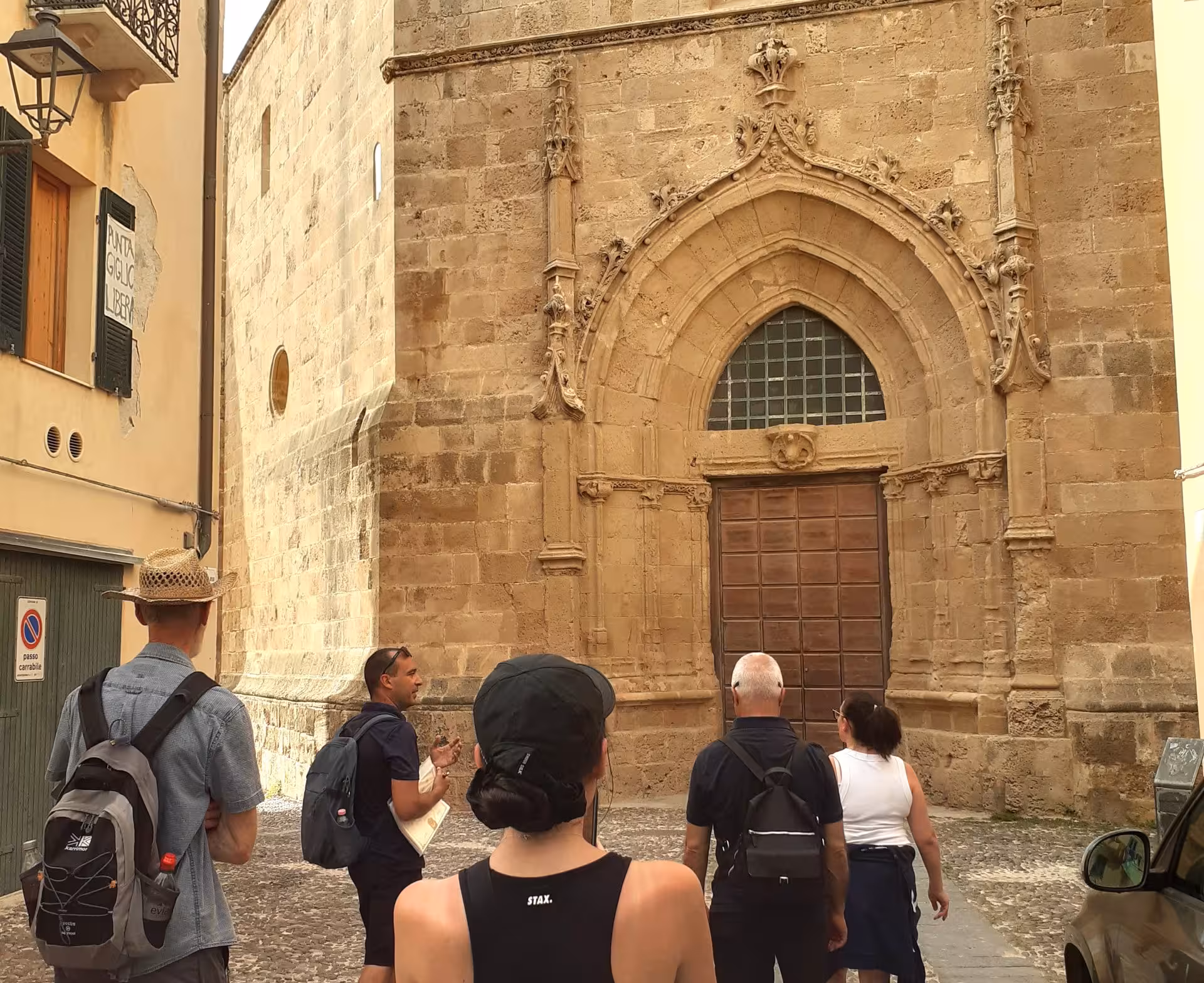 Tourists explore Alghero's historic center with a guide, admiring the intricate architecture of an ancient stone building.