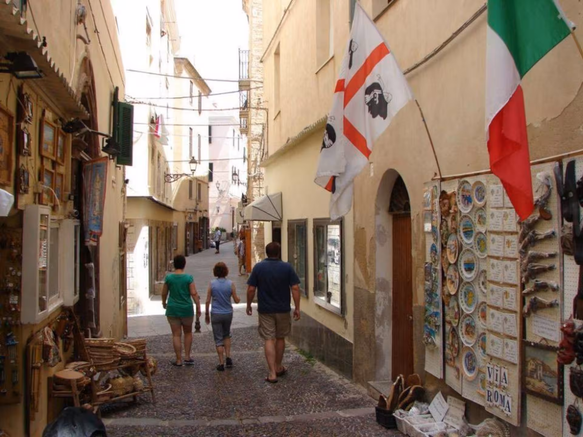 Tourists strolling through a narrow street with flags and local shops in Alghero's historic center.