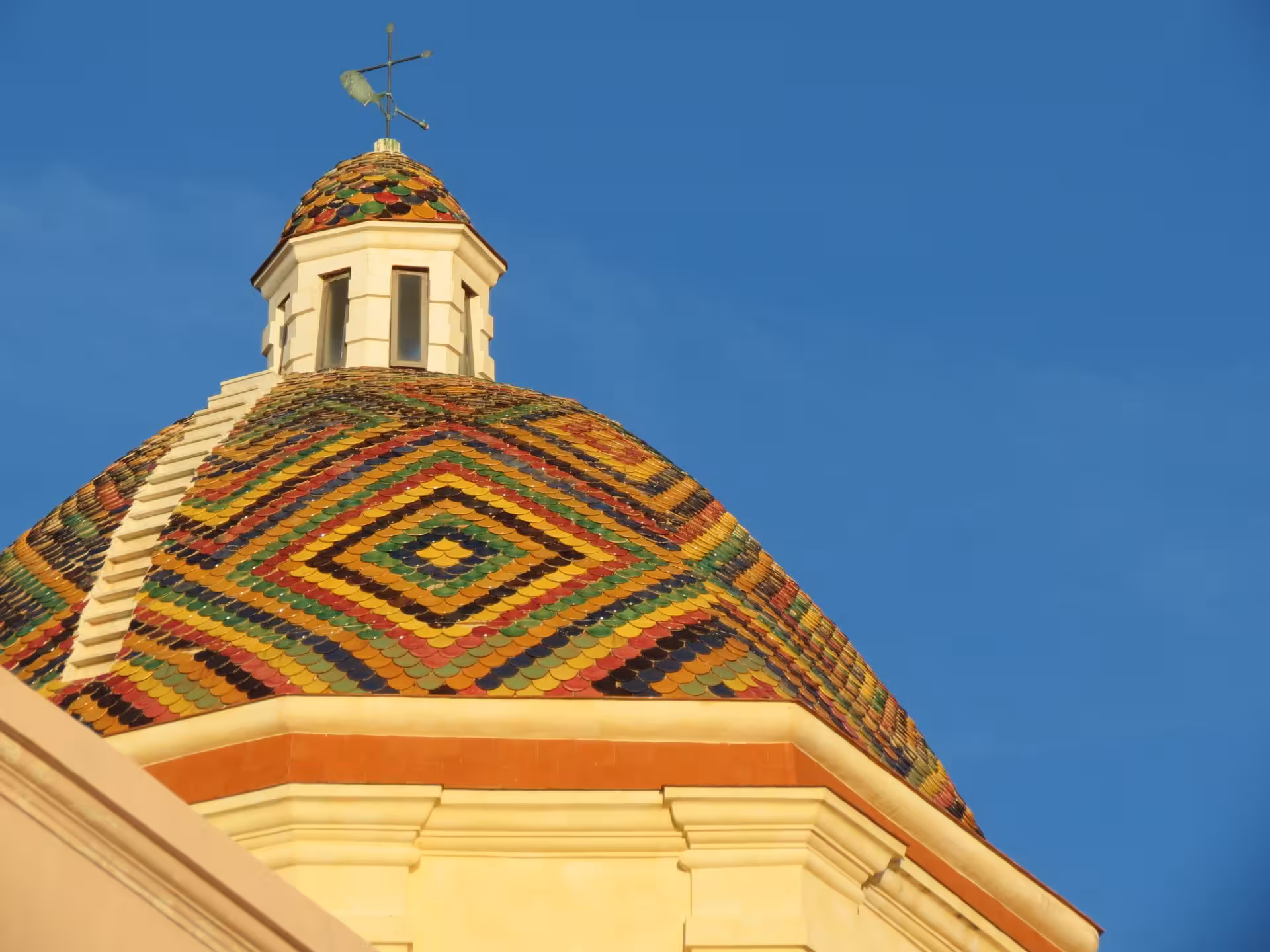 Vibrant tiled dome of Alghero's historic church under a clear blue sky, showcasing Sardinian architectural charm.