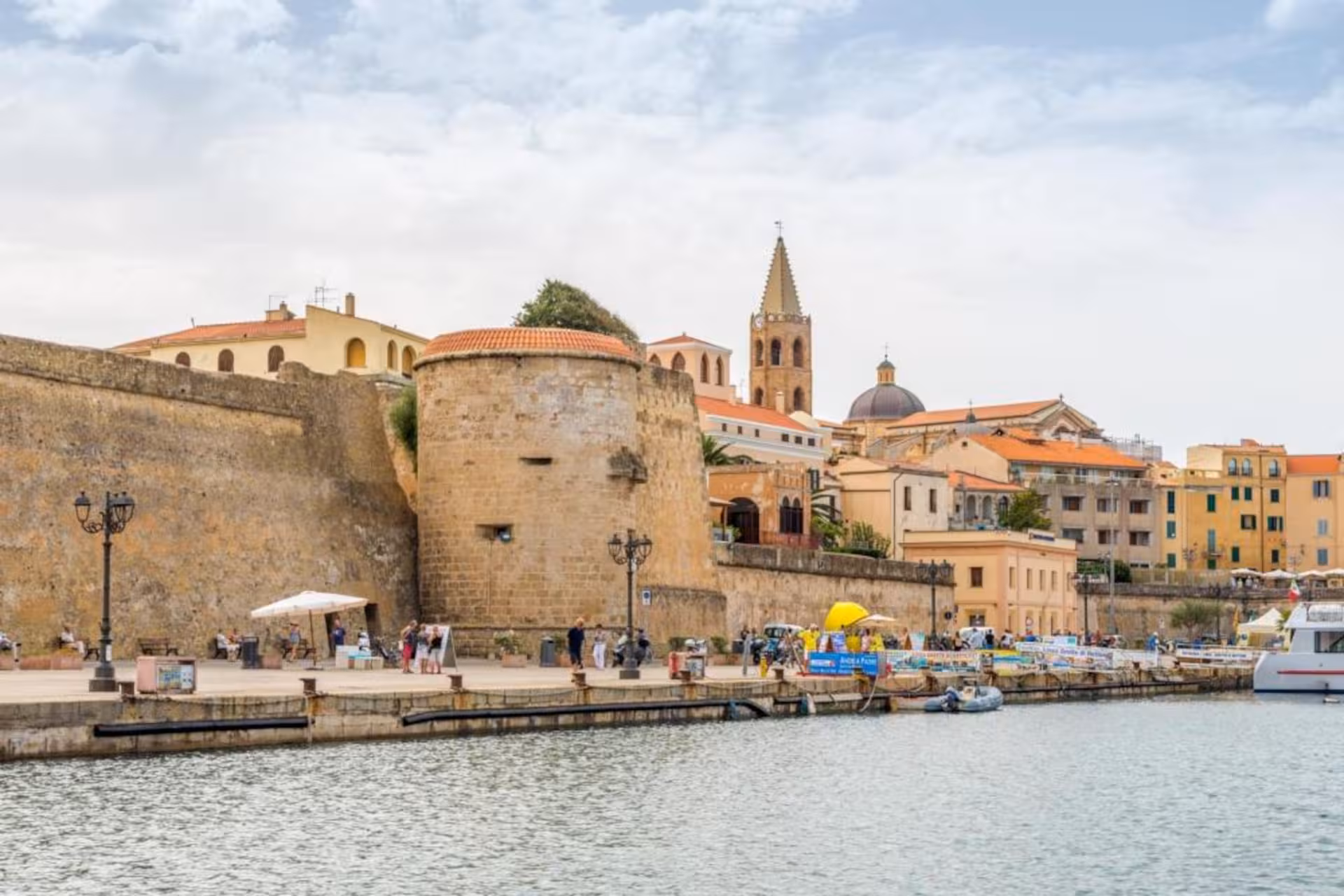 Scenic view of Alghero's historic waterfront with ancient walls and bell towers under a cloudy sky.