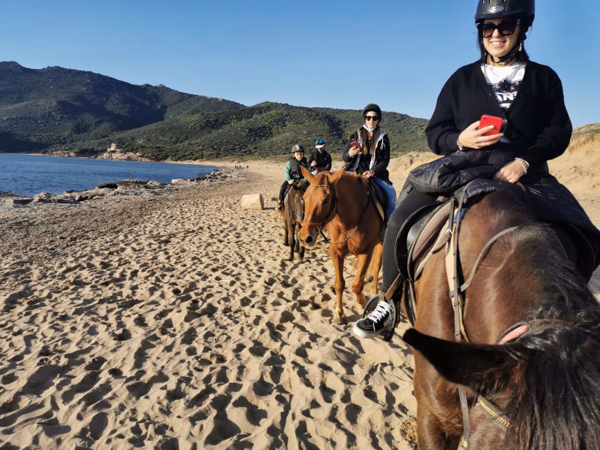 Smiling riders enjoying a sunny horseback tour along Porto Ferro's sandy shores in Alghero, capturing memories.
