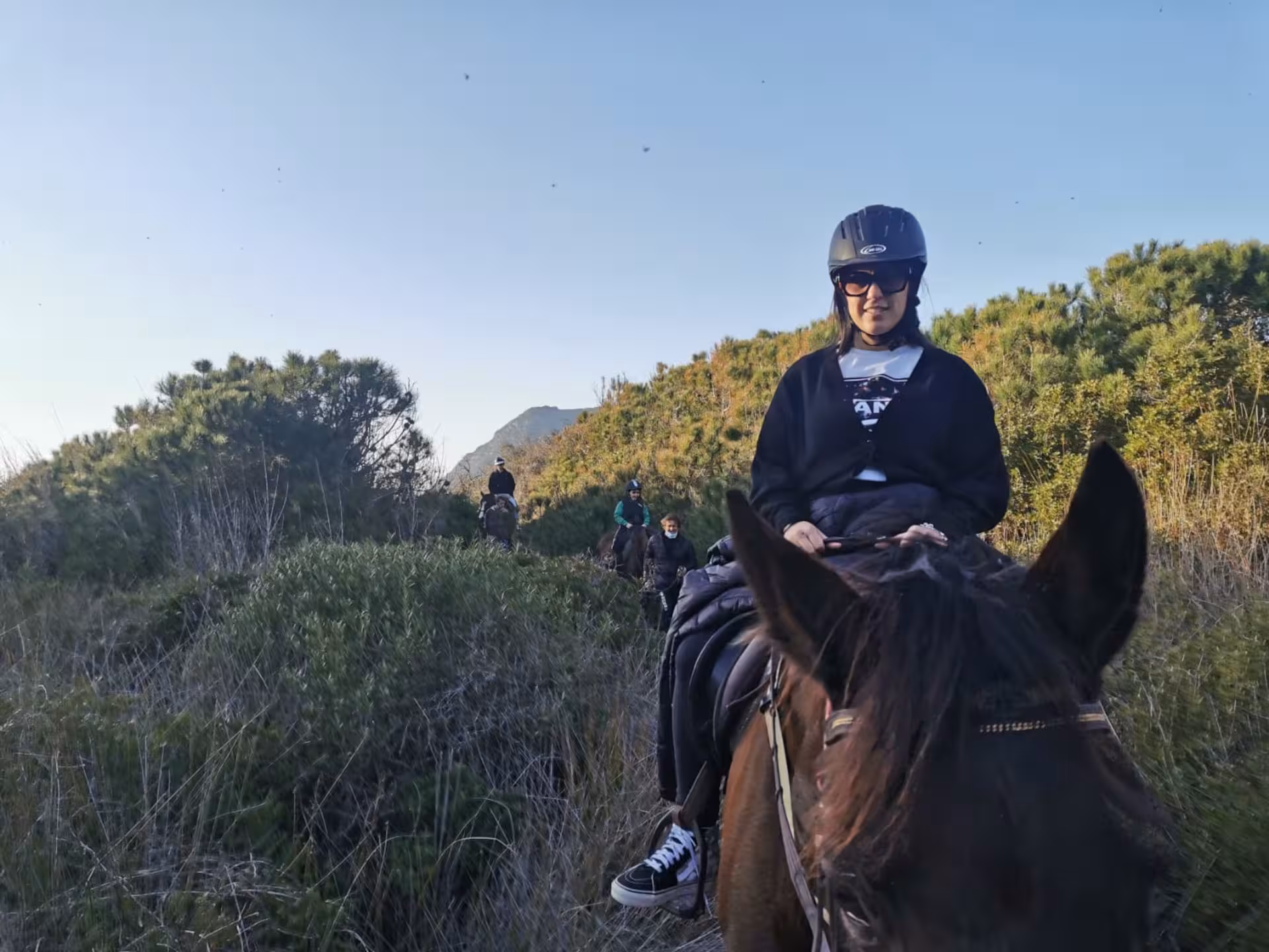 Horseback riders navigate lush trails in Alghero's Porto Ferro, surrounded by vibrant greenery and clear skies.