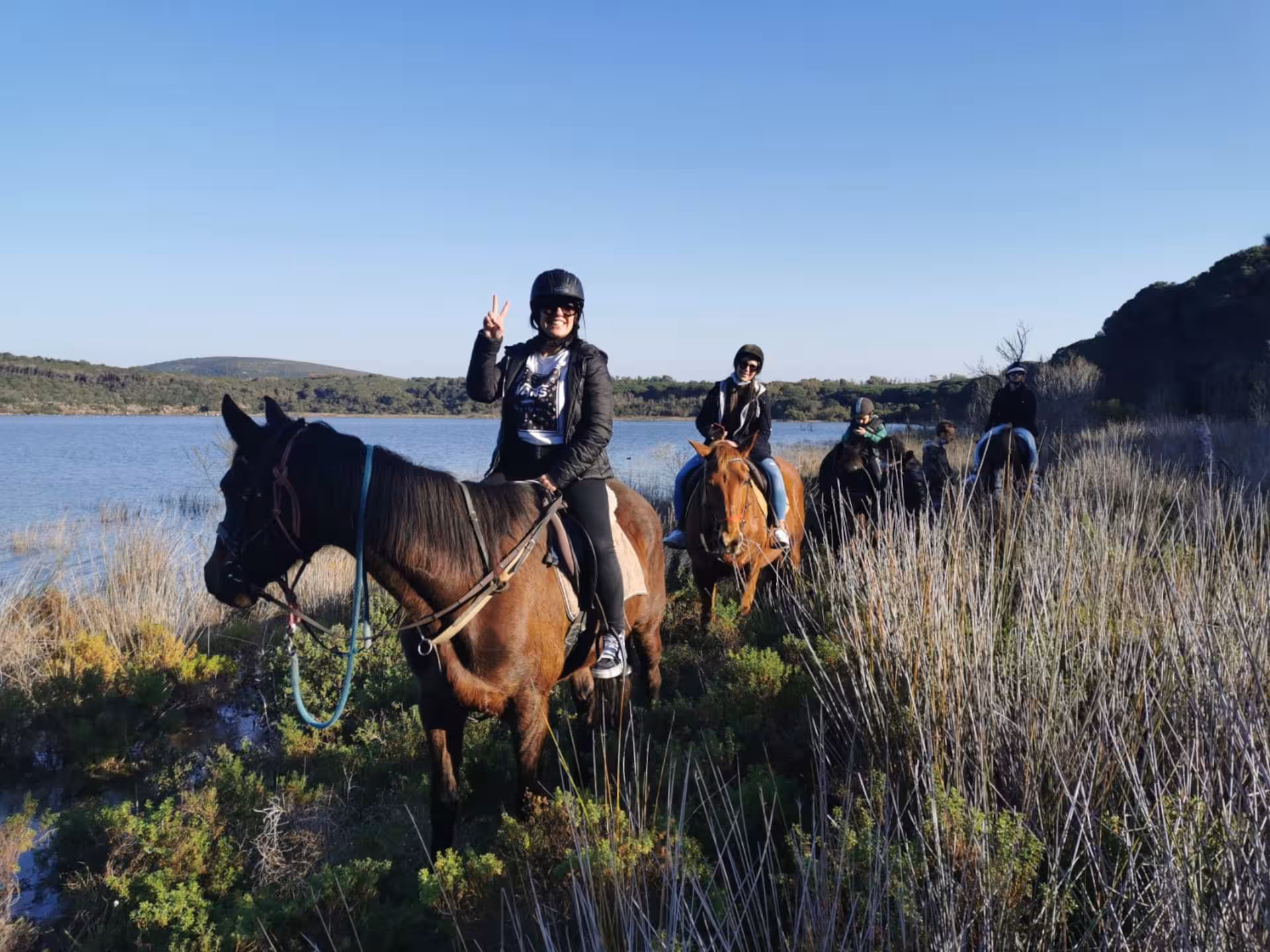 Riders enjoy a scenic horseback ride along Lake Baratz in Alghero, surrounded by lush vegetation and clear skies.