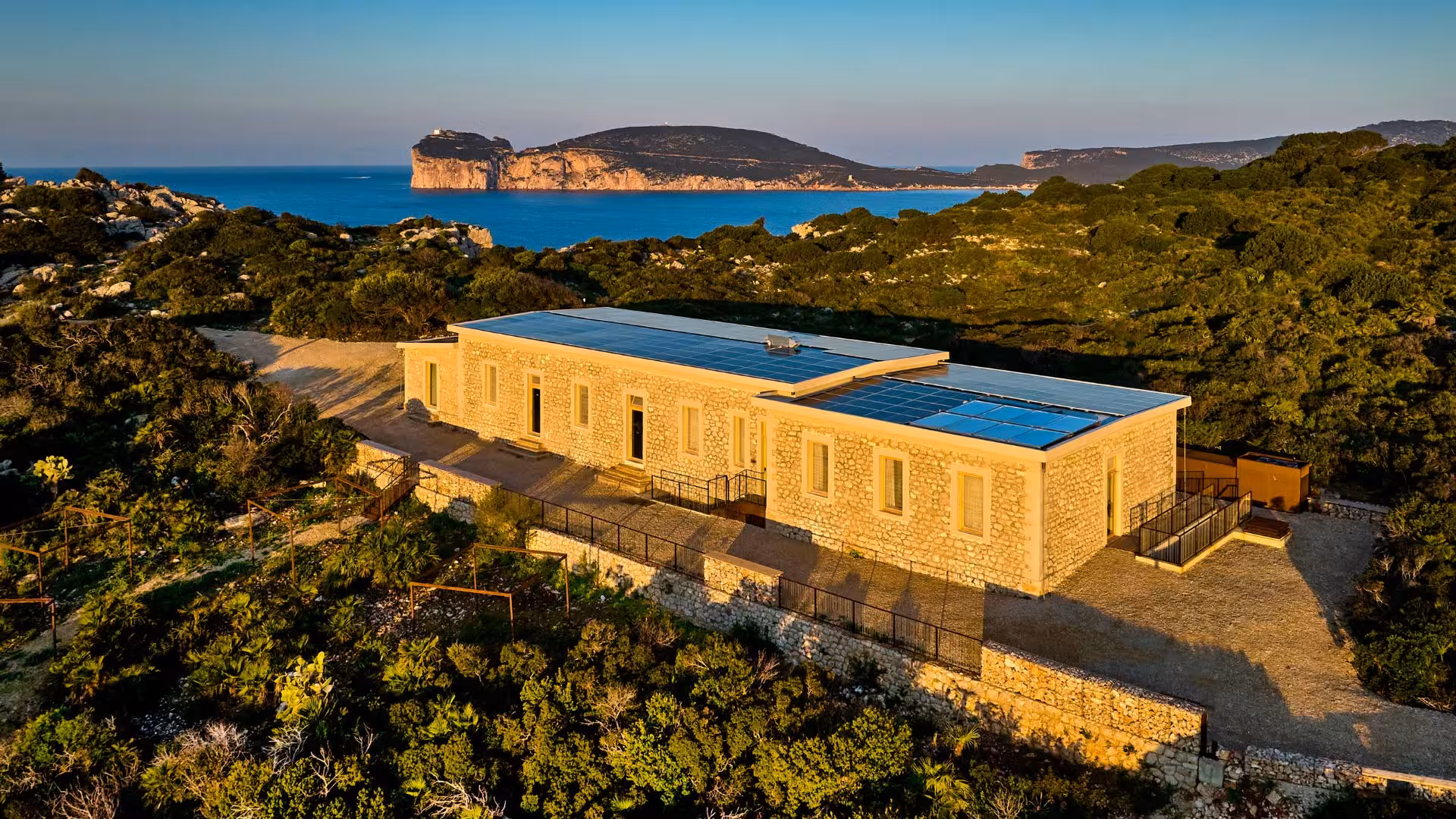 Stone building amidst lush greenery in Porto Conte Park, Alghero, with sea and cliffs in the background, ideal for trekking.
