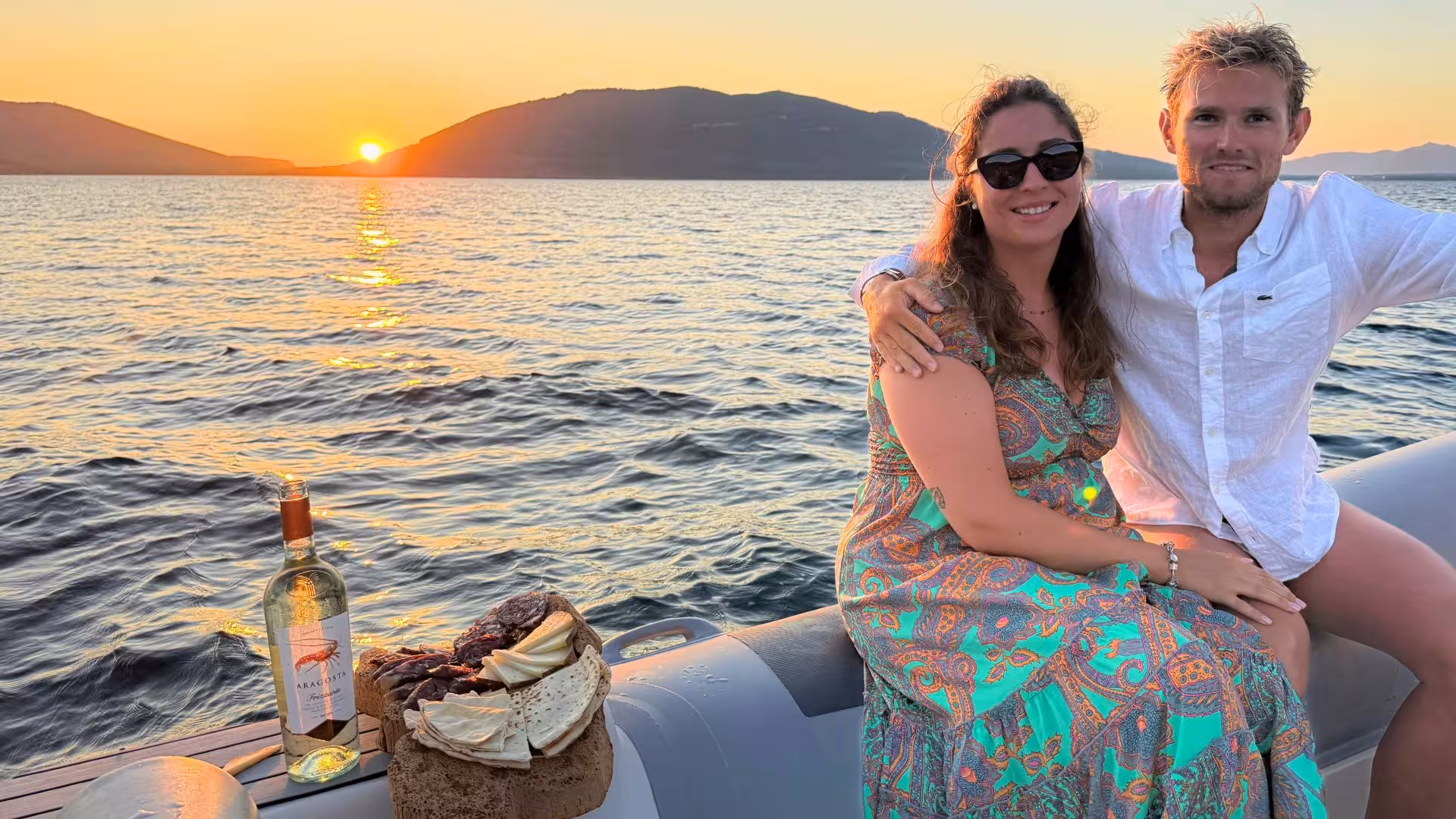 Couple enjoying a romantic sunset on a dinghy in Alghero with wine and snacks, perfect for a private tour experience.