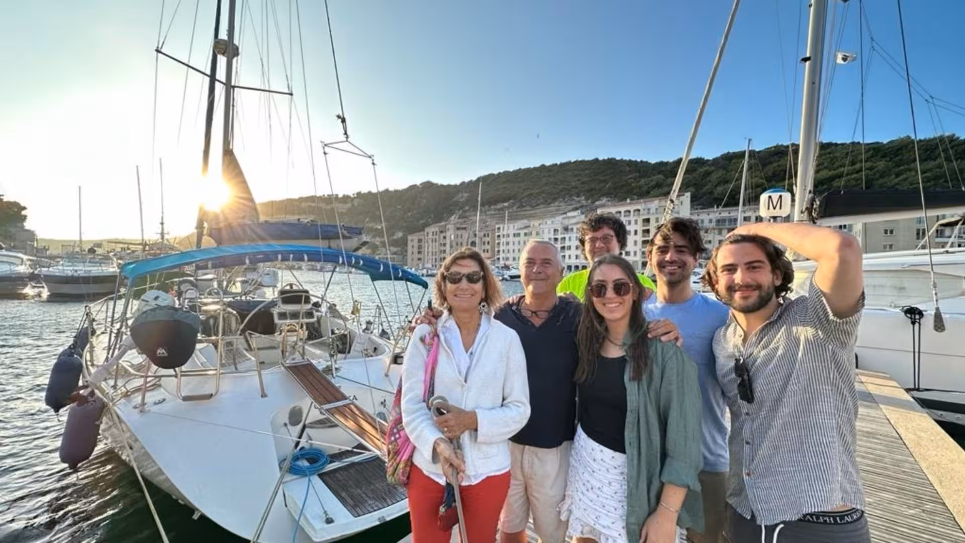 Group of people smiling on a sailboat in Alghero harbor, enjoying a picturesque sailing adventure.